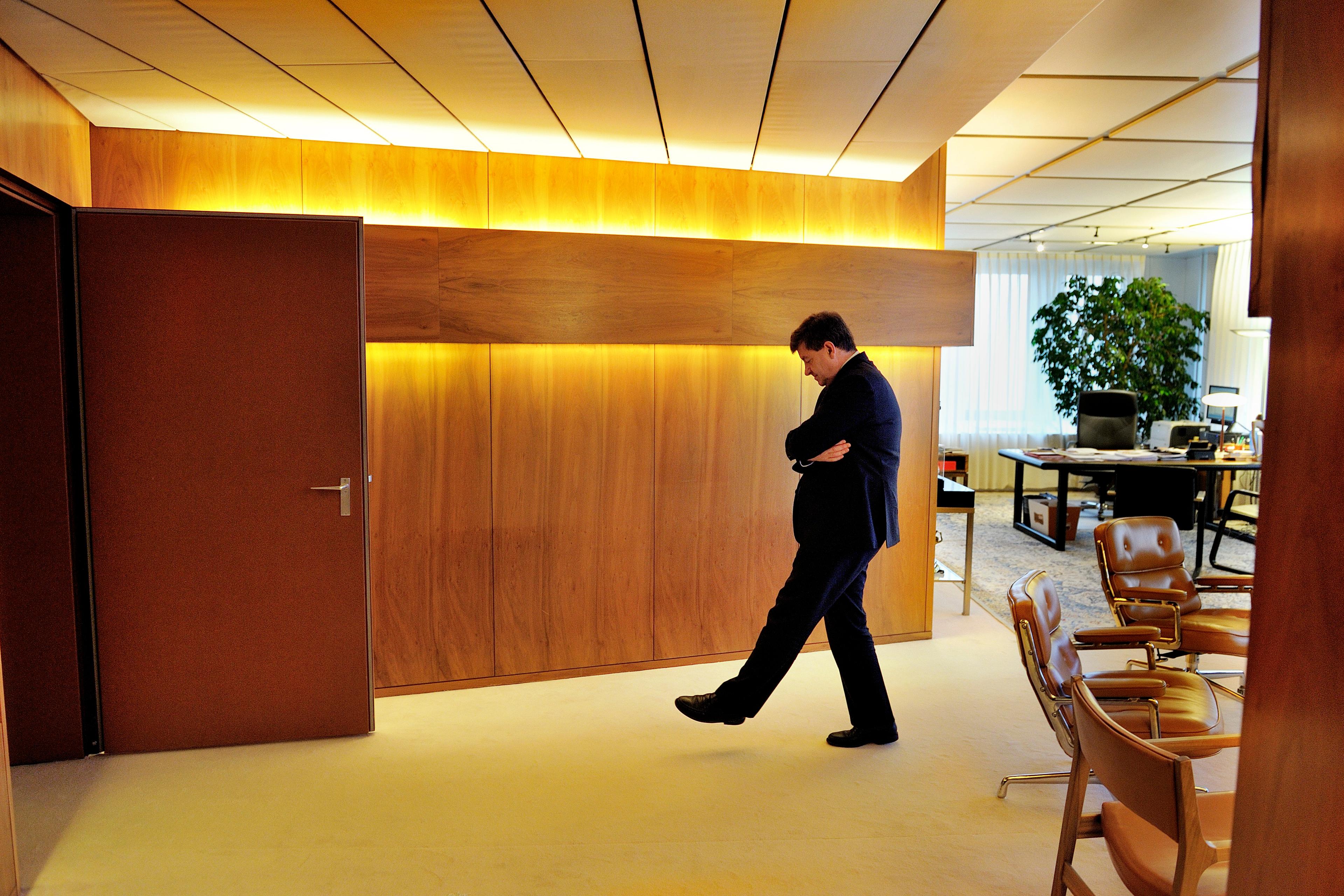 Photo of a man in an office walking pensively, with wooden walls and brown chairs around.
