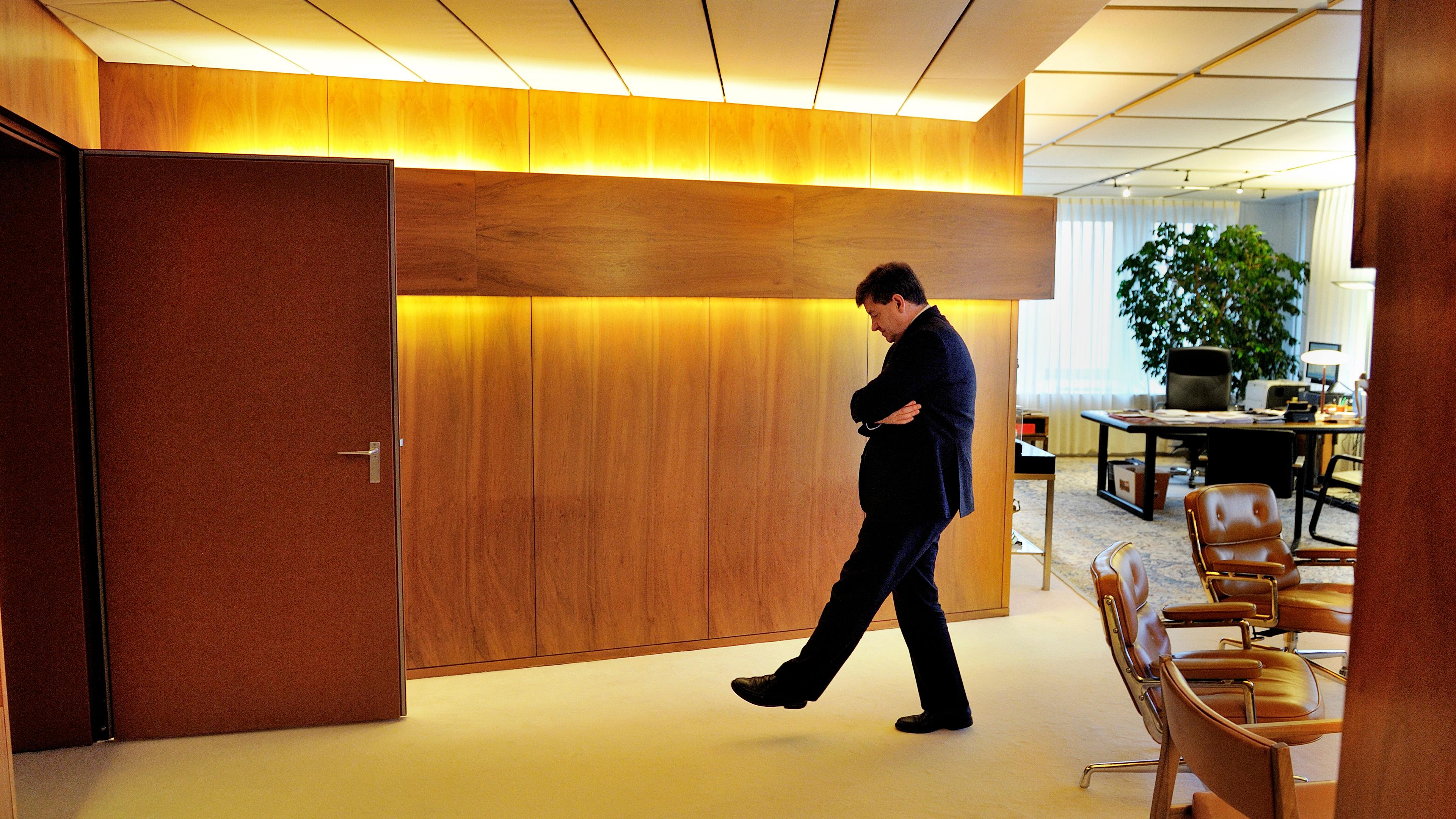 Photo of a man in an office walking pensively, with wooden walls and brown chairs around.