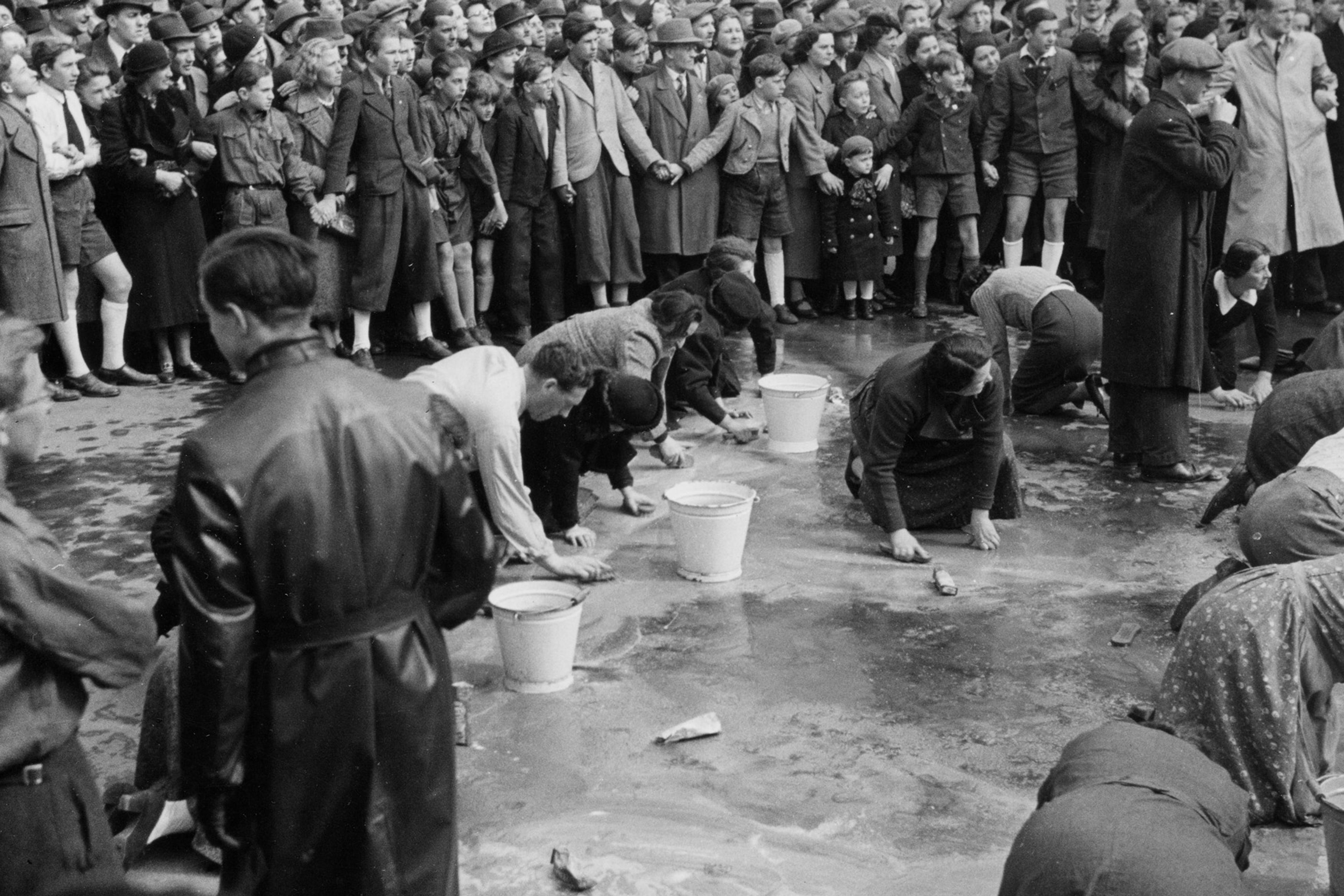 Black and white photo of people scrubbing a street while a large crowd of onlookers stands watching.