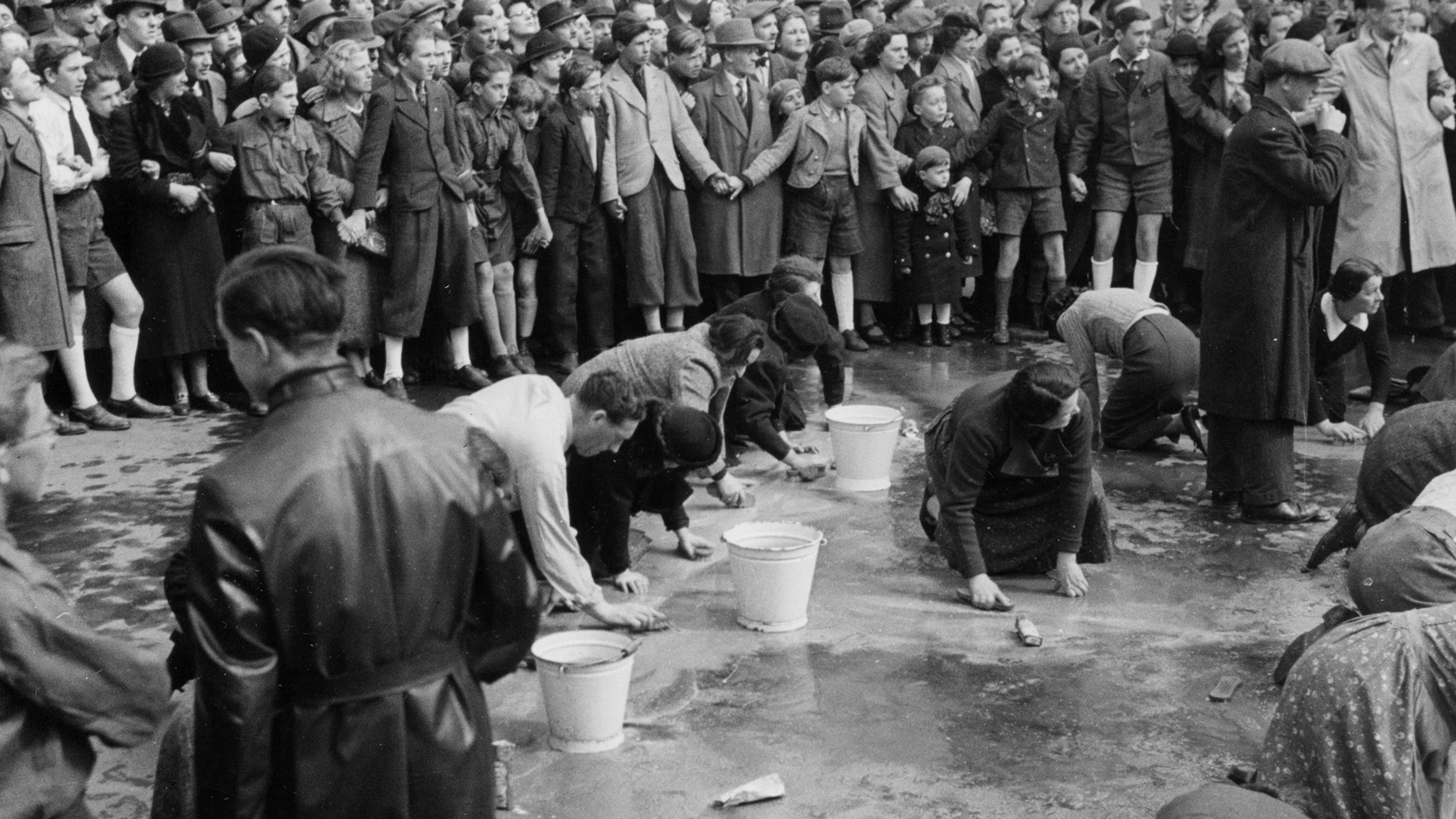 Black and white photo of people scrubbing a street while a large crowd of onlookers stands watching.