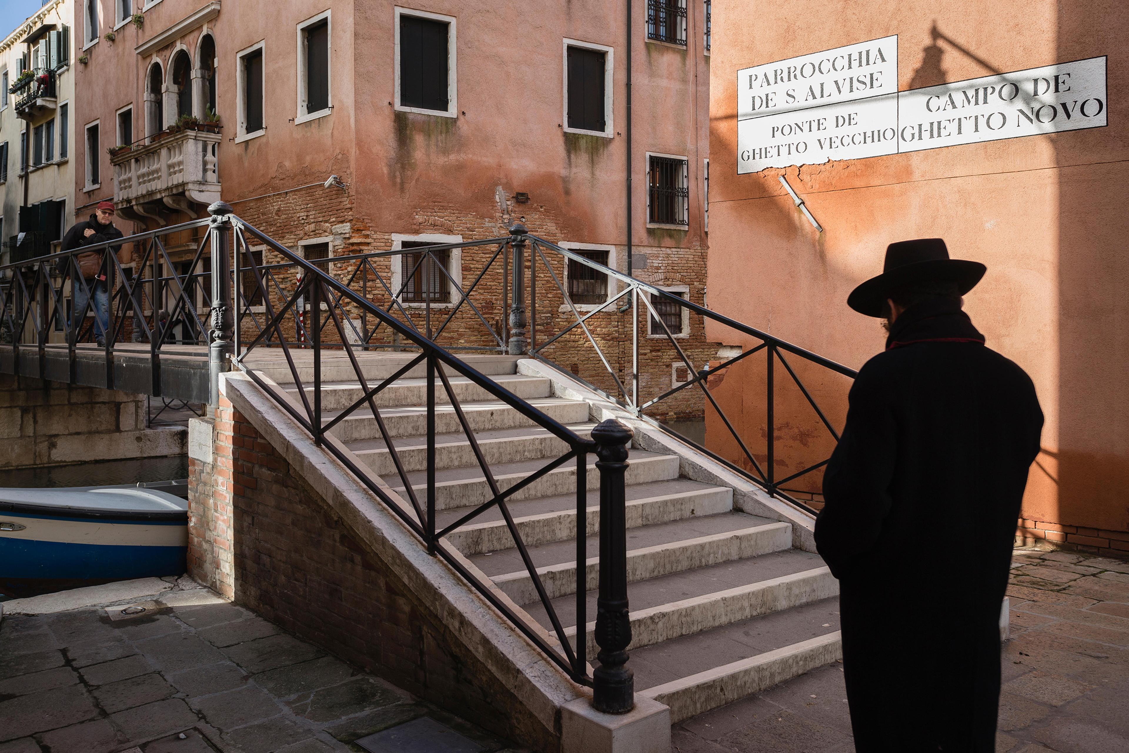 A person in black clothing and a hat by a bridge and stairs in a historic area, with buildings and signs in the background.