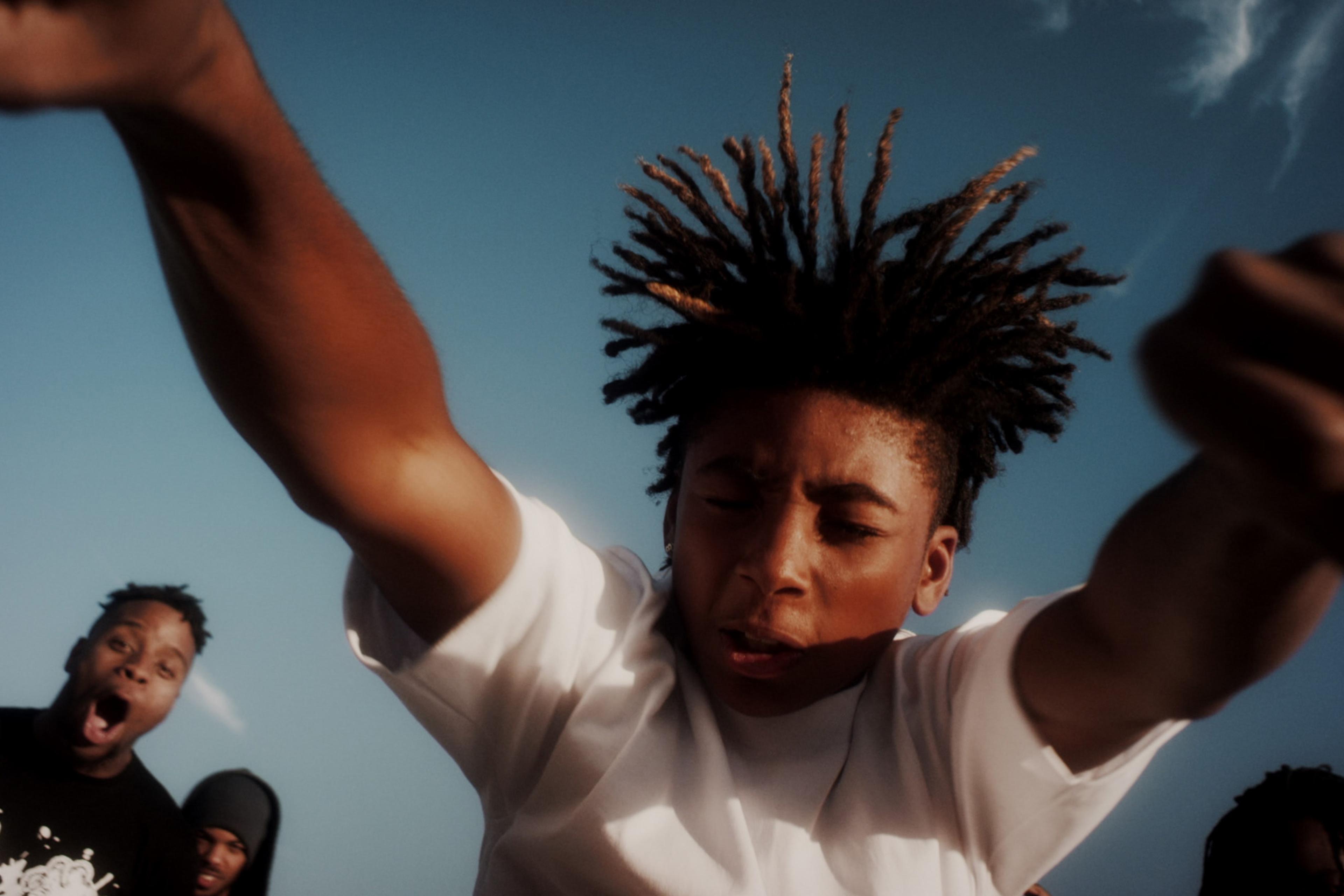 A young person dancing with arms extended and hair flying, seen from below, with sky background. Two other excited people watch.