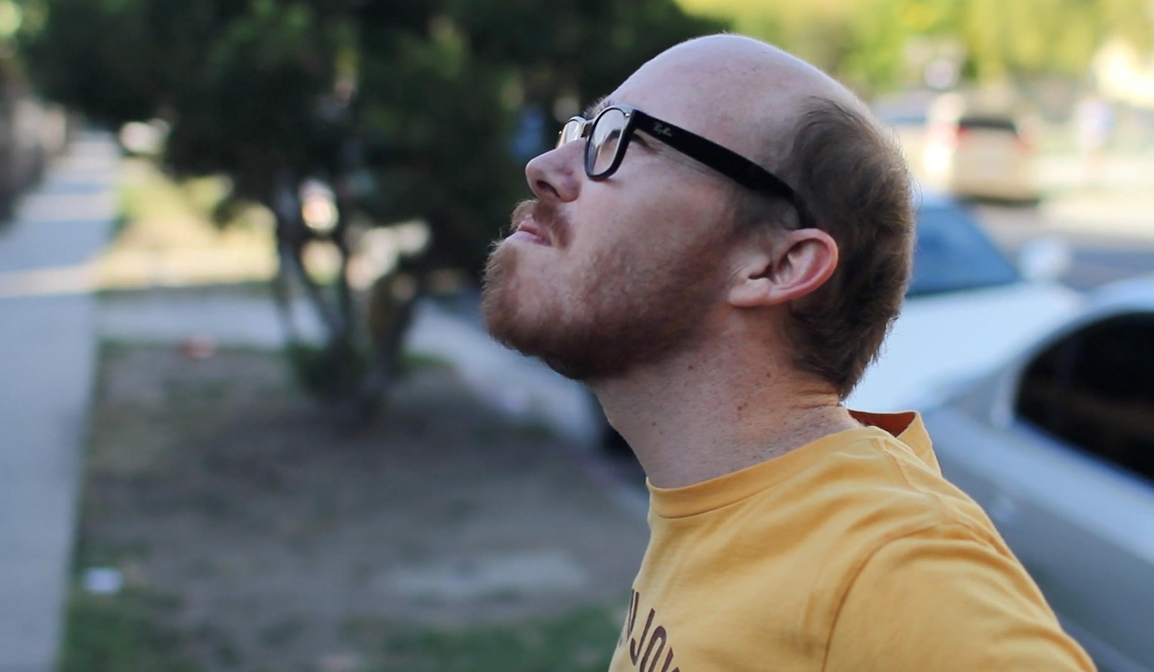 Bearded man with glasses and a yellow T-shirt looking up, standing outside near a pavement and trees.