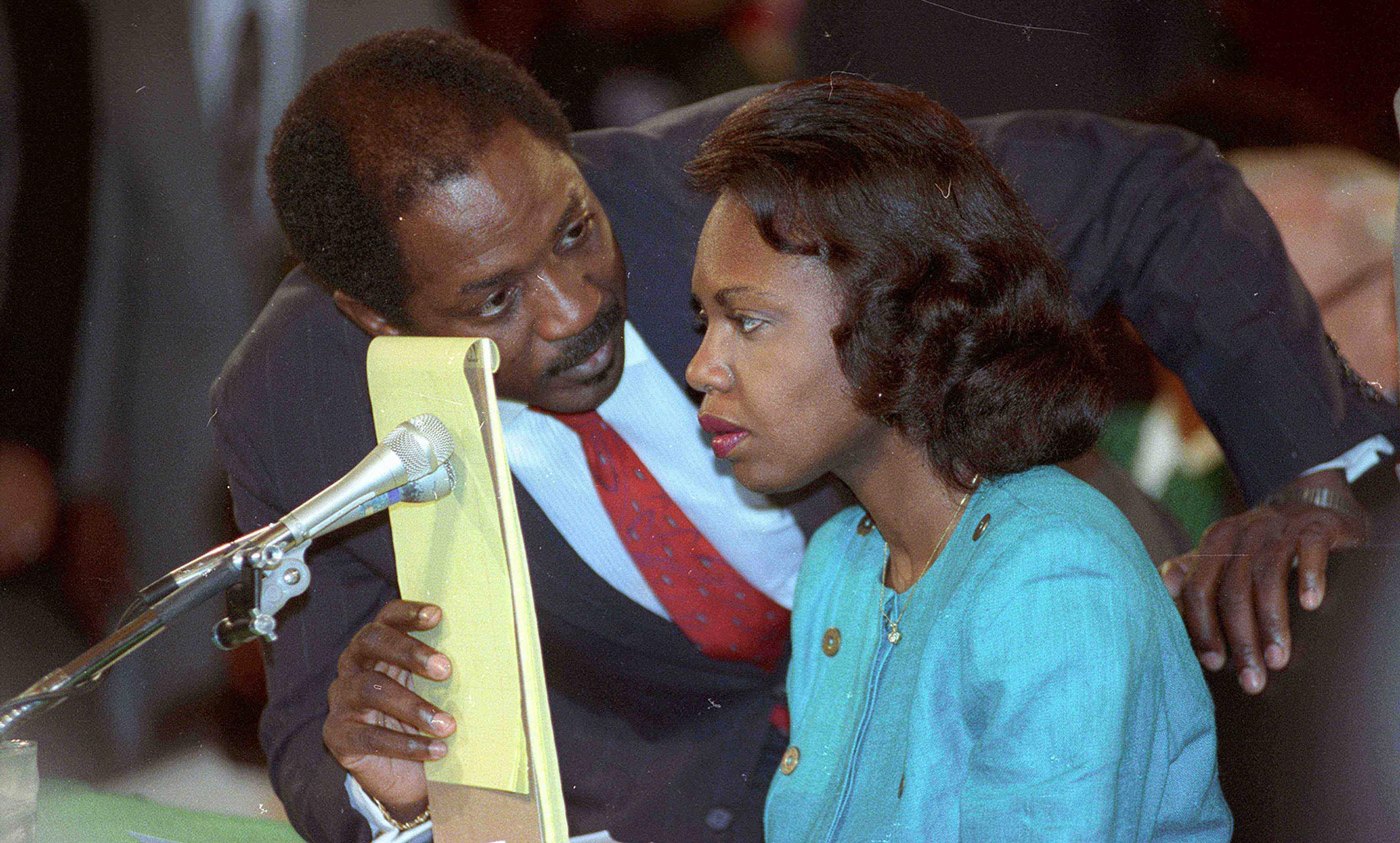 Photo of two people conversing, the man is holding papers near a microphone while the woman listens intently with a sombre expression.