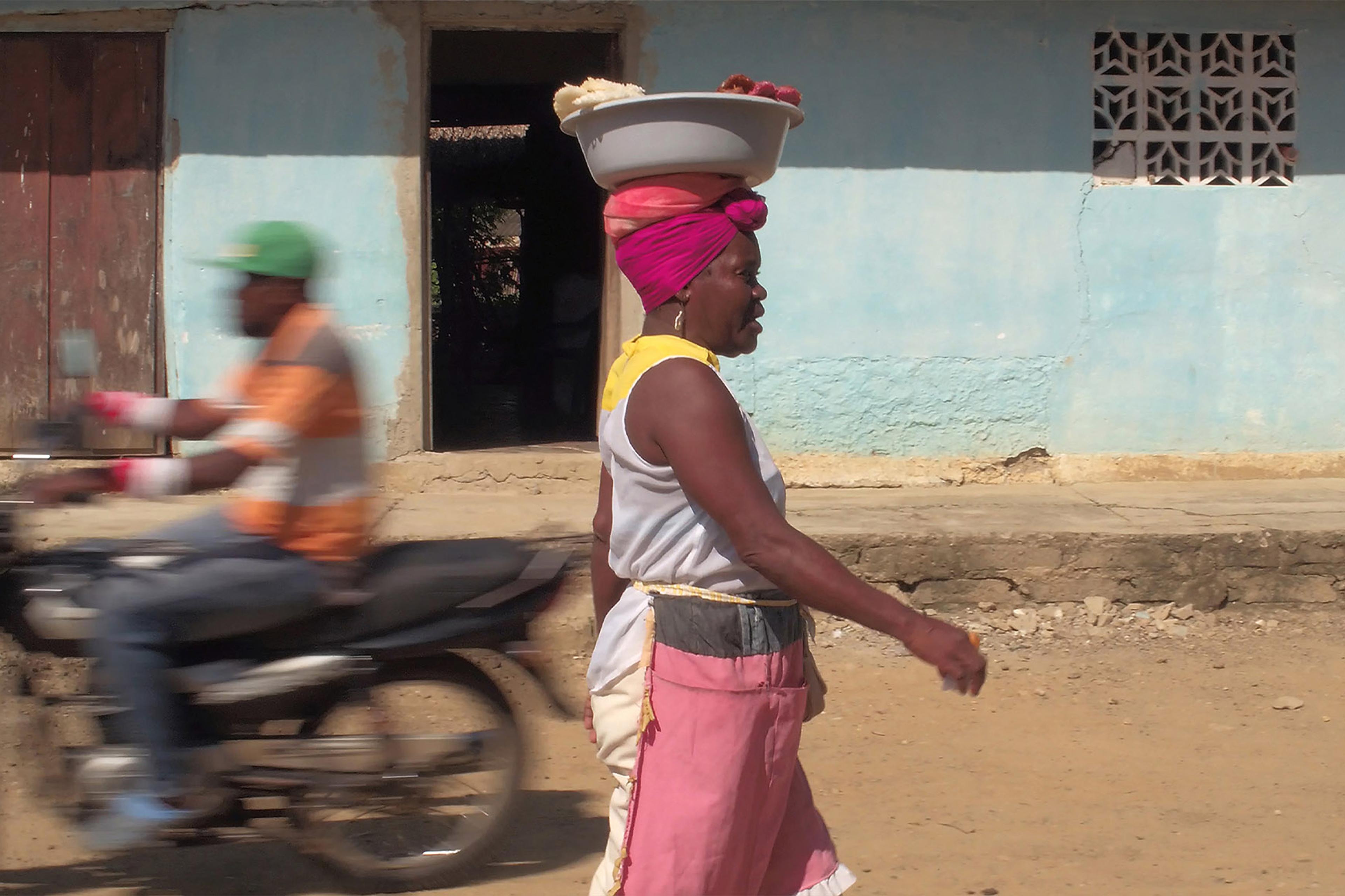 A Black woman balancing a basin on her head walking on a dirt road past a moving motorcycle and a blue building.