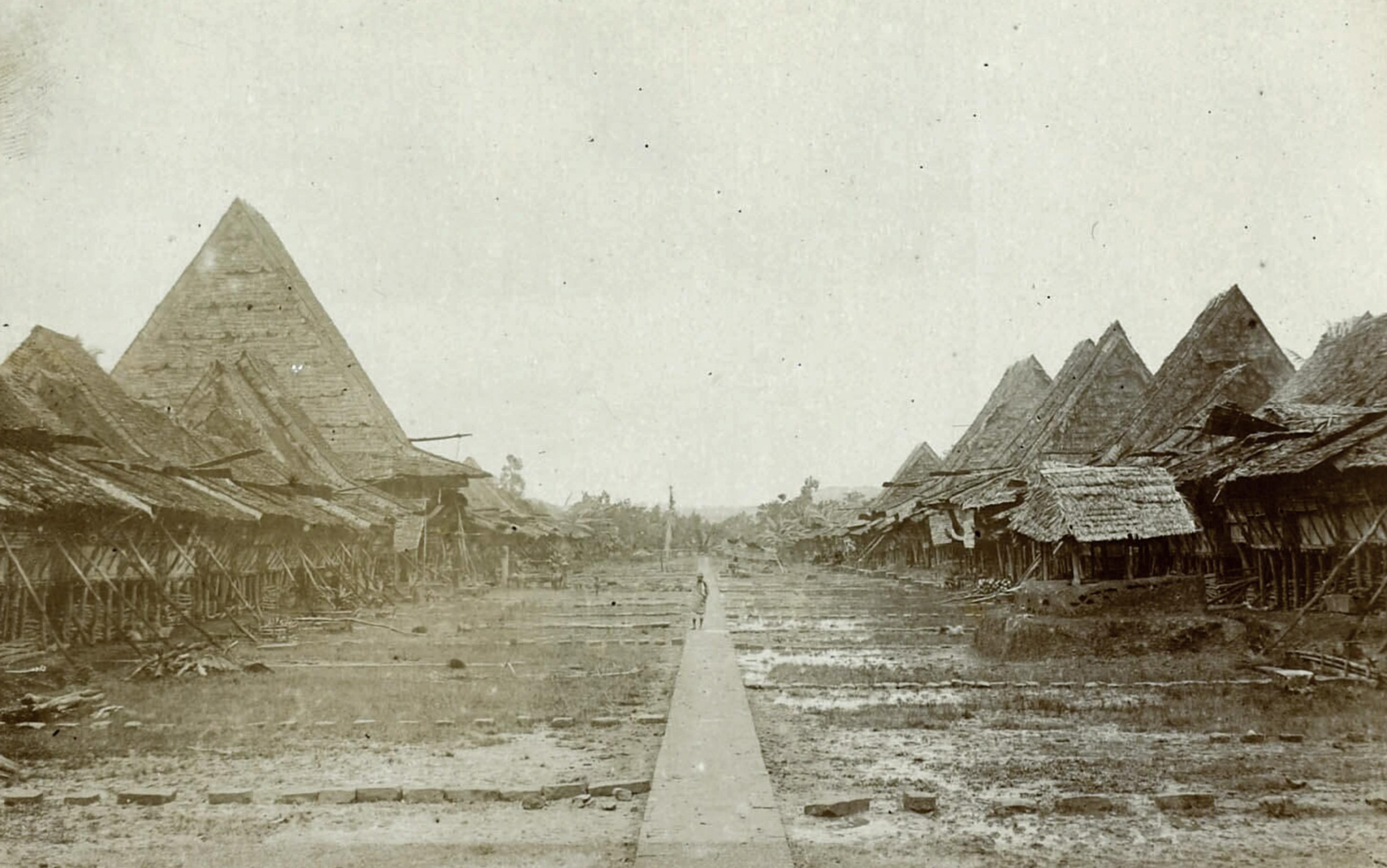 Black-and-white photo of traditional thatched huts lining a dirt road in a village, with a lone figure walking in the centre.