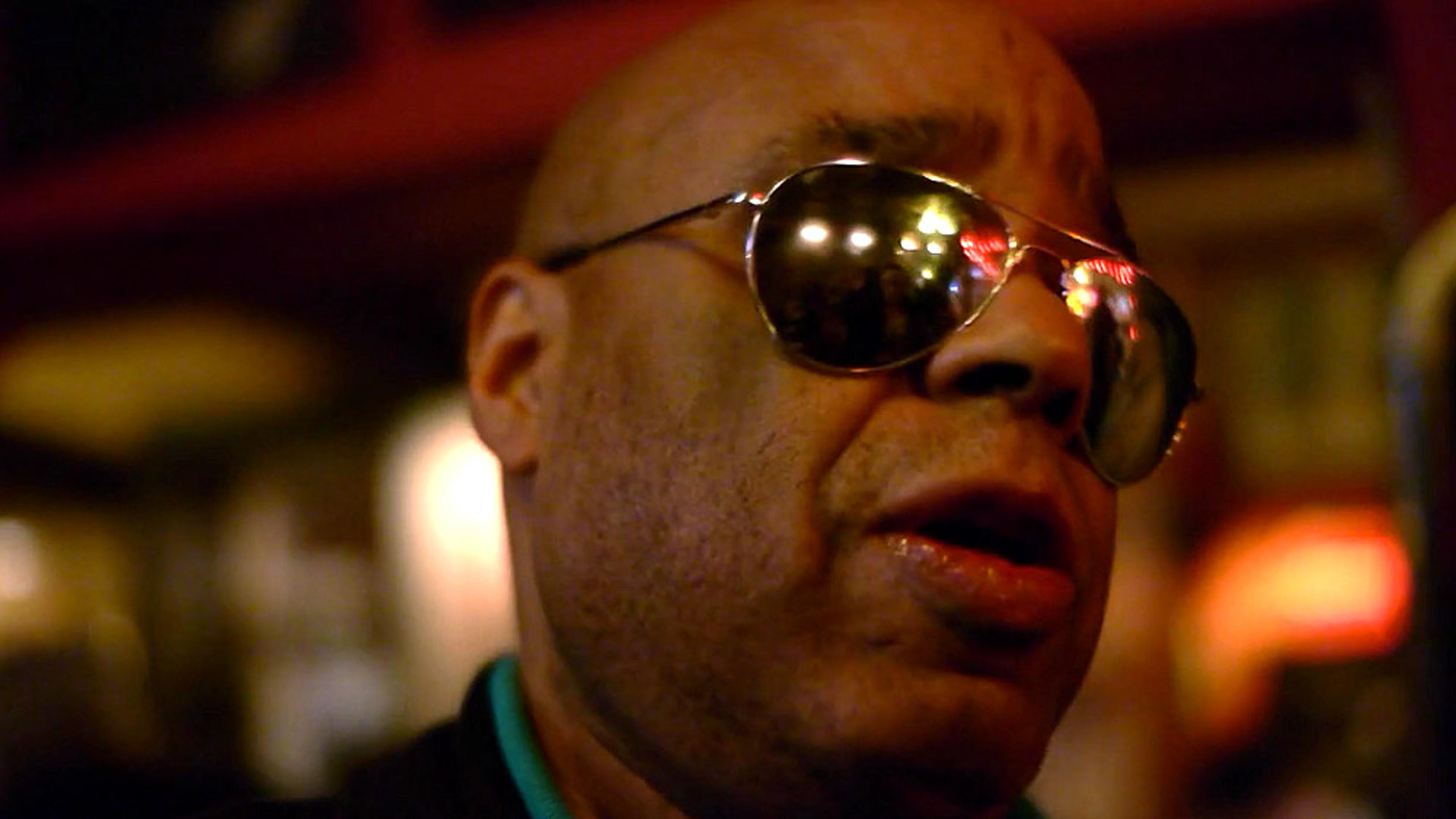 A Black man wearing sunglasses looking to the side in a dimly lit environment with reflections in his glasses.