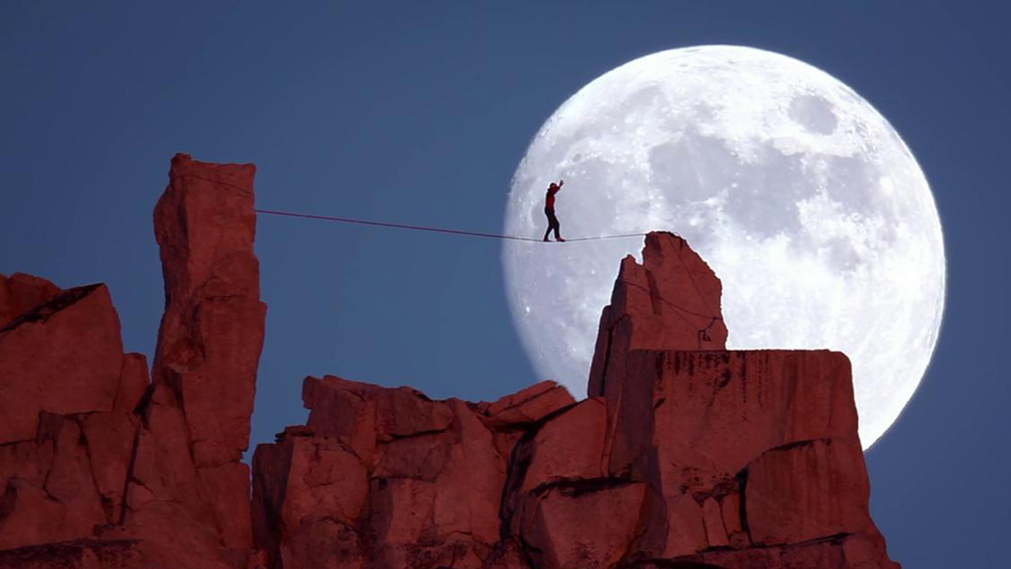 A person walking on a tightrope between rock formations with a large full moon in the background.