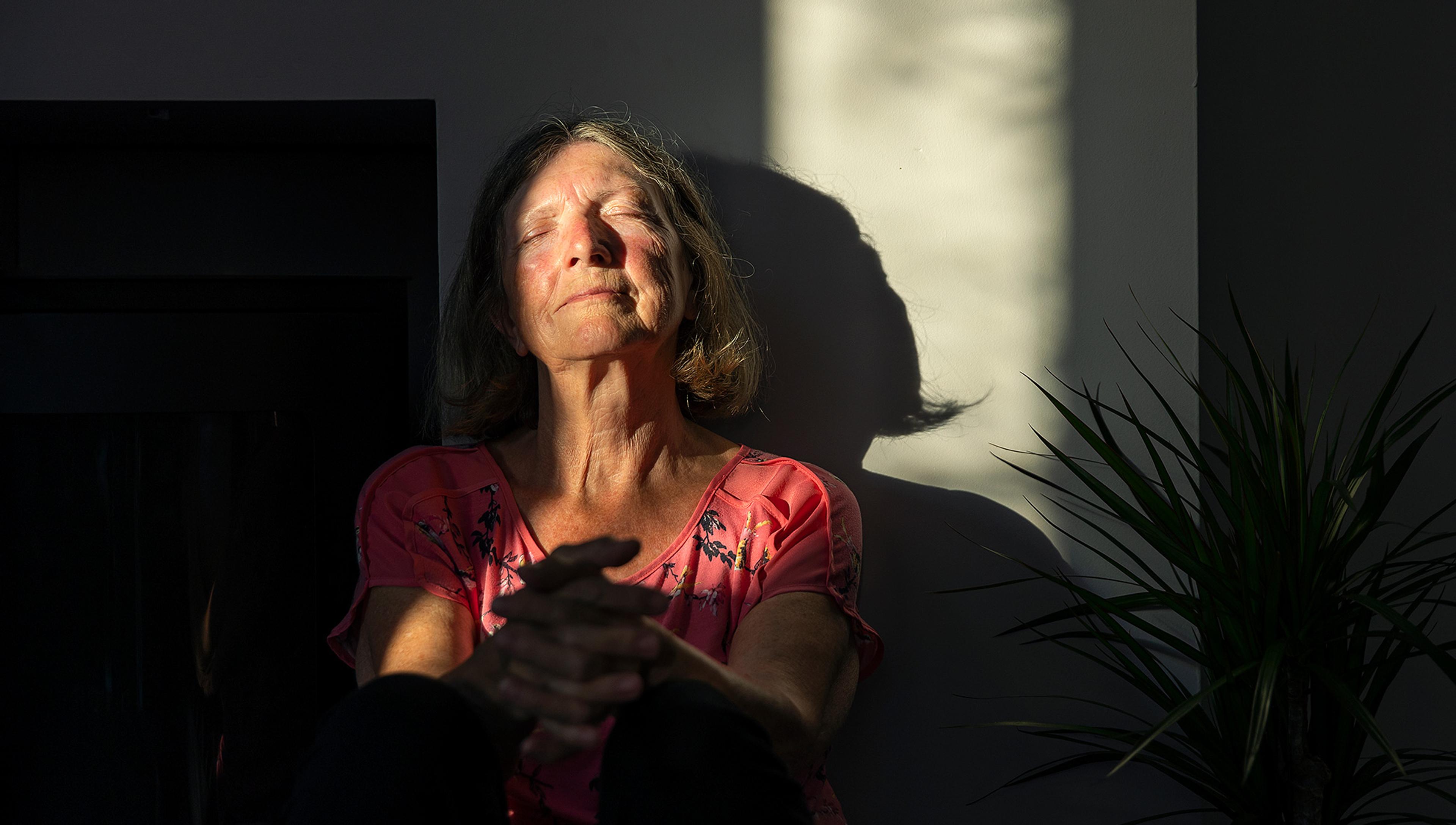 A woman in sunlight with eyes closed, wearing a pink top sitting by a shadowy wall and green plant.