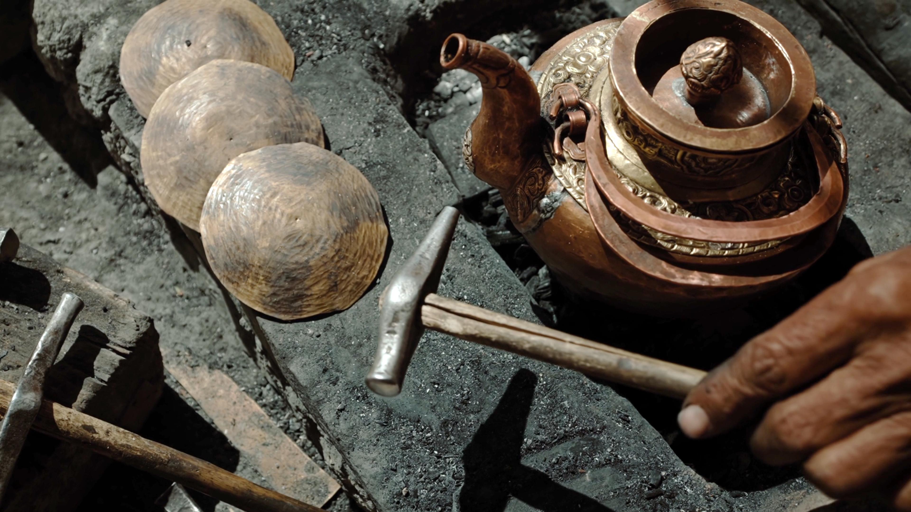 Close-up of a hammer, metal pot, and three copper spheres on a stone surface, with a hand holding the hammer.