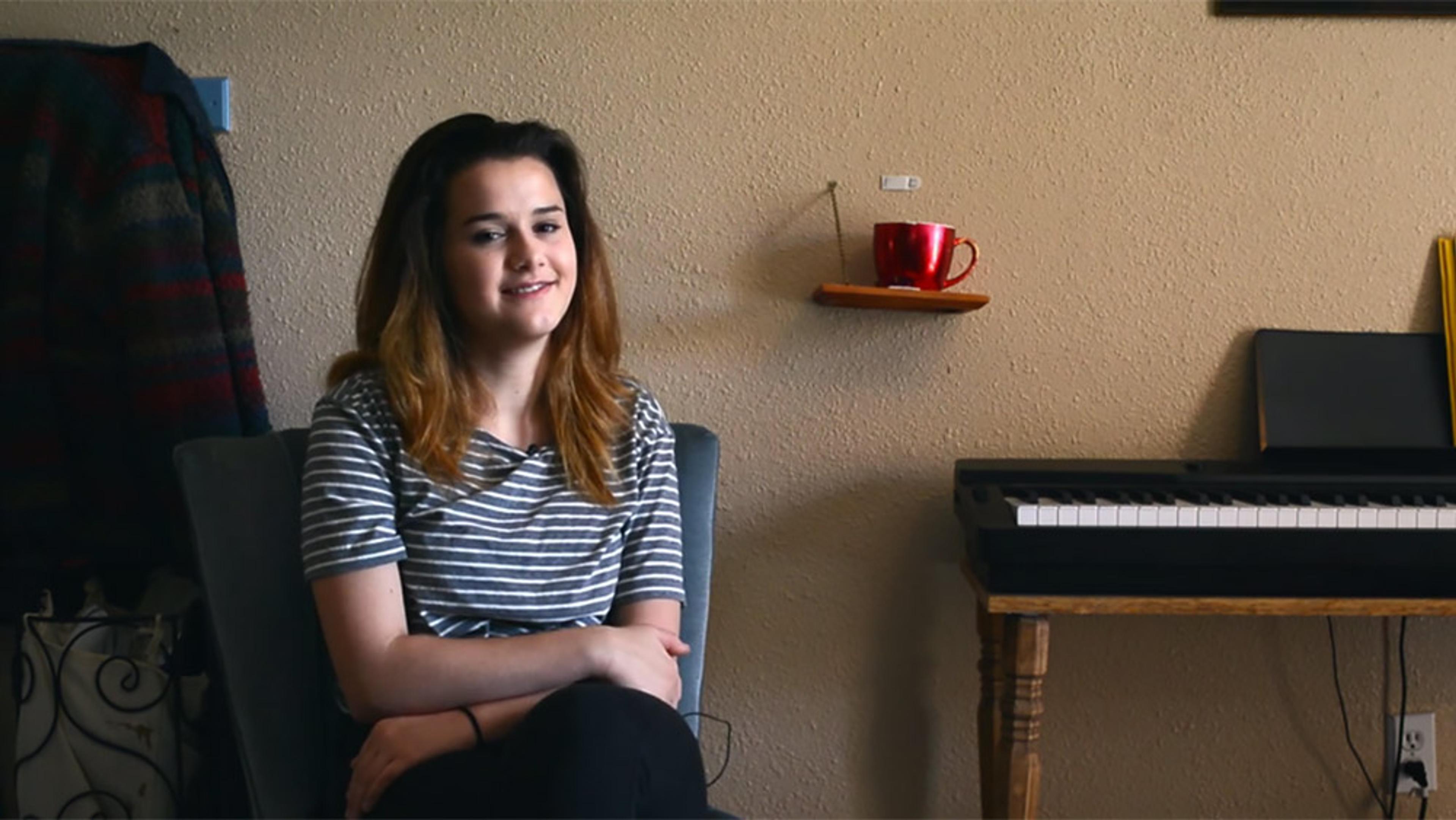 A smiling young woman sitting in a chair in a room with a keyboard, a red mug on a small wall shelf, and a coat on a hanger.