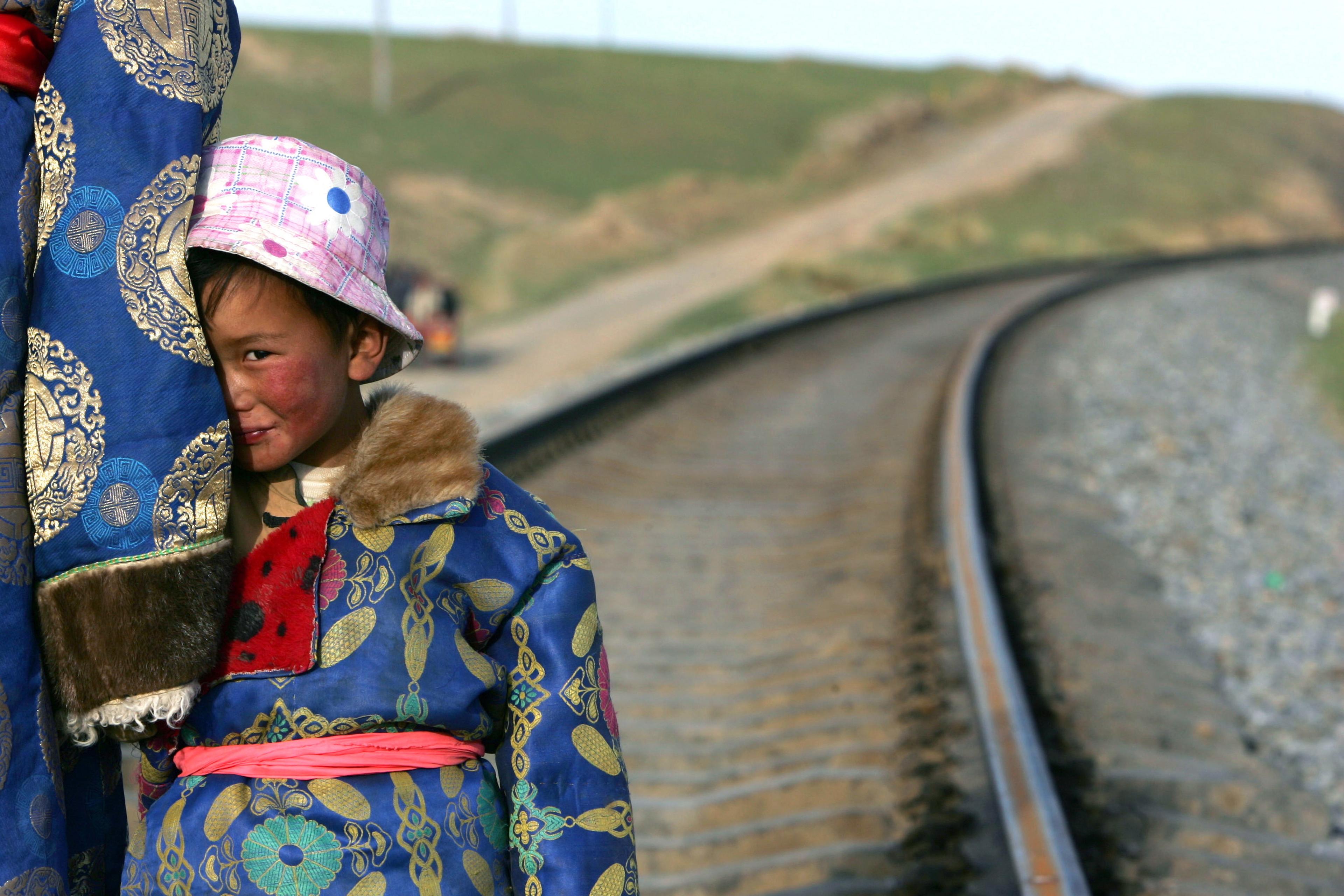 A photo of a child in a patterned blue coat and pink hat standing by railway tracks looking to the side.