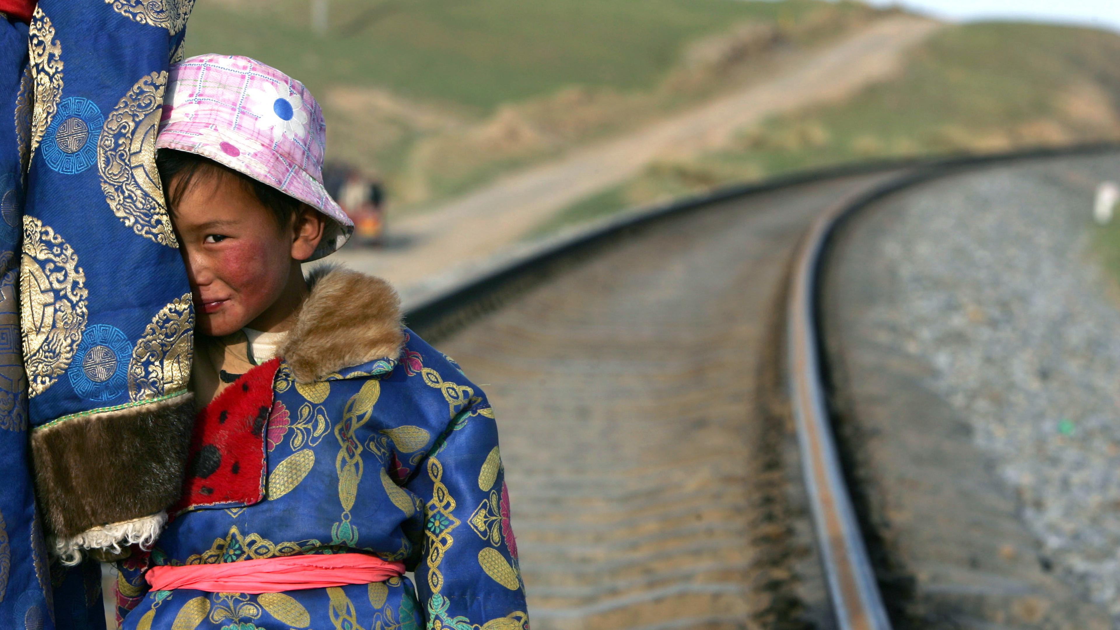A photo of a child in a patterned blue coat and pink hat standing by railway tracks looking to the side.
