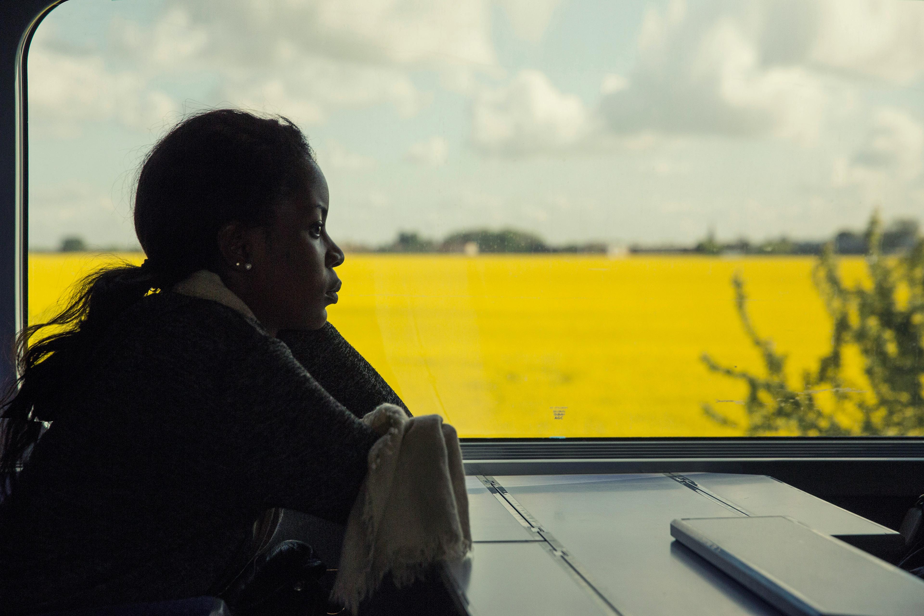 A person gazing out a train window at a yellow field and cloudy sky, with a table and a closed laptop in view.