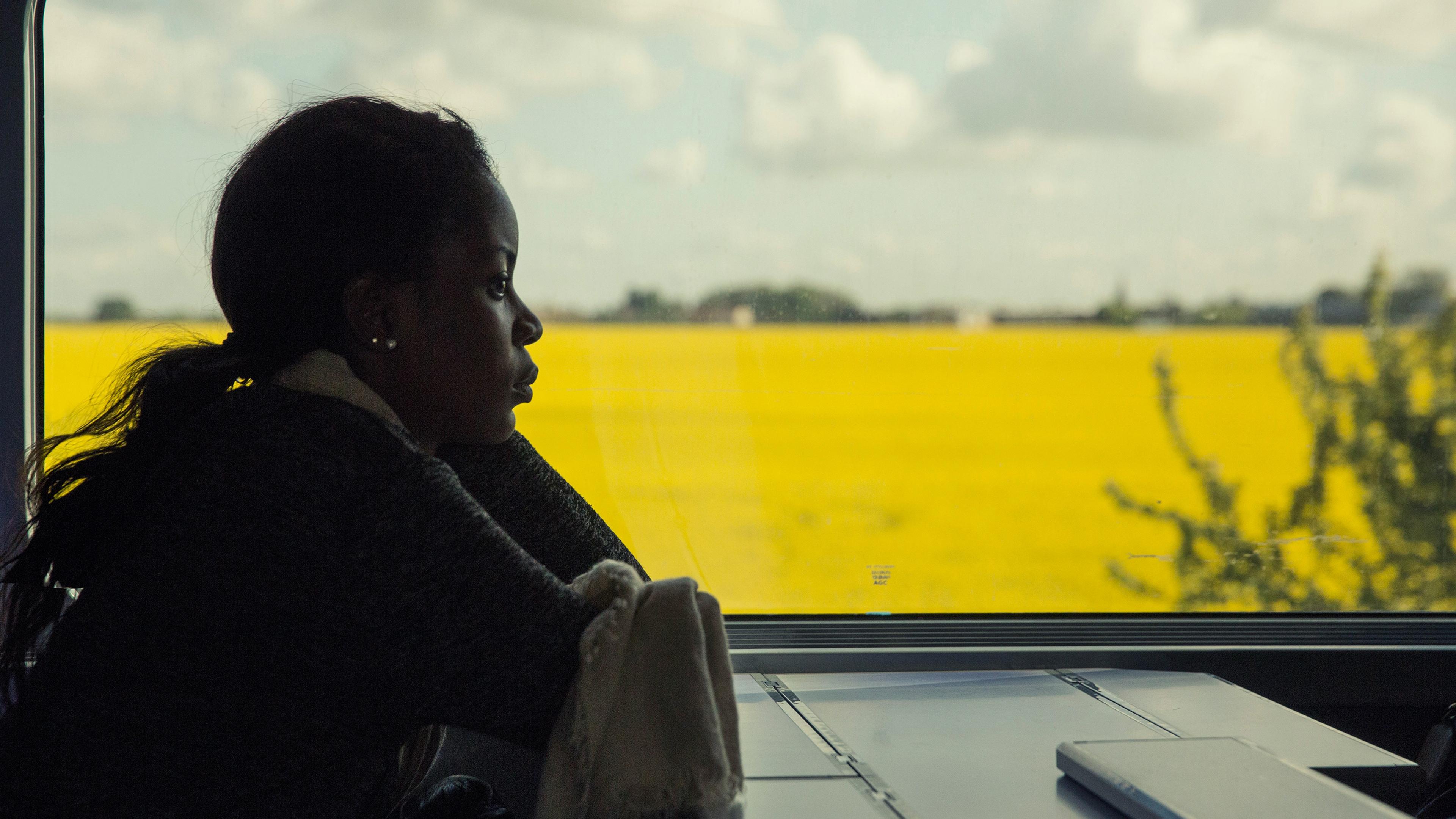A person gazing out a train window at a yellow field and cloudy sky, with a table and a closed laptop in view.