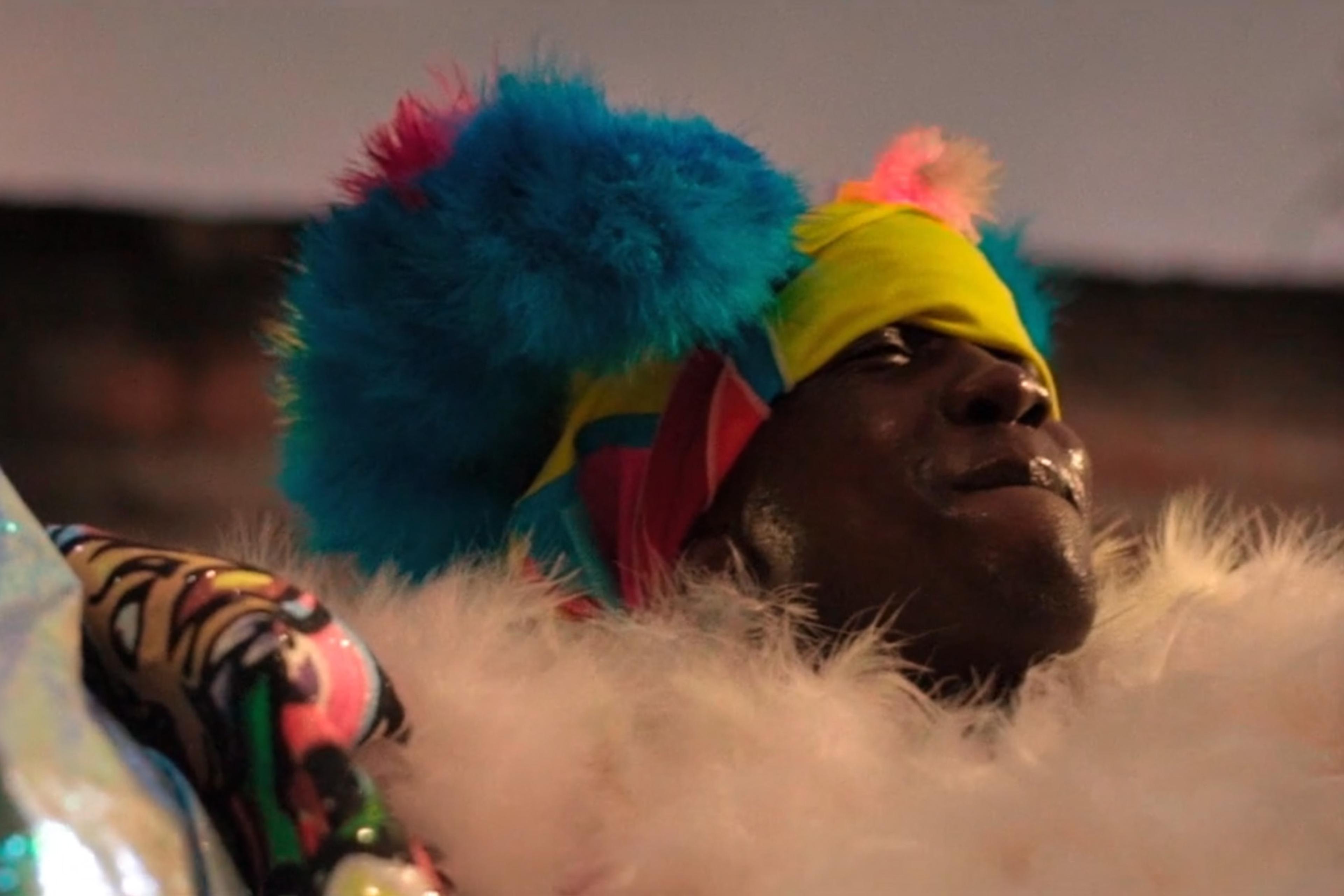 A person in colourful costume with a yellow and blue headdress, blue fluffy accessories and a white feather boa.