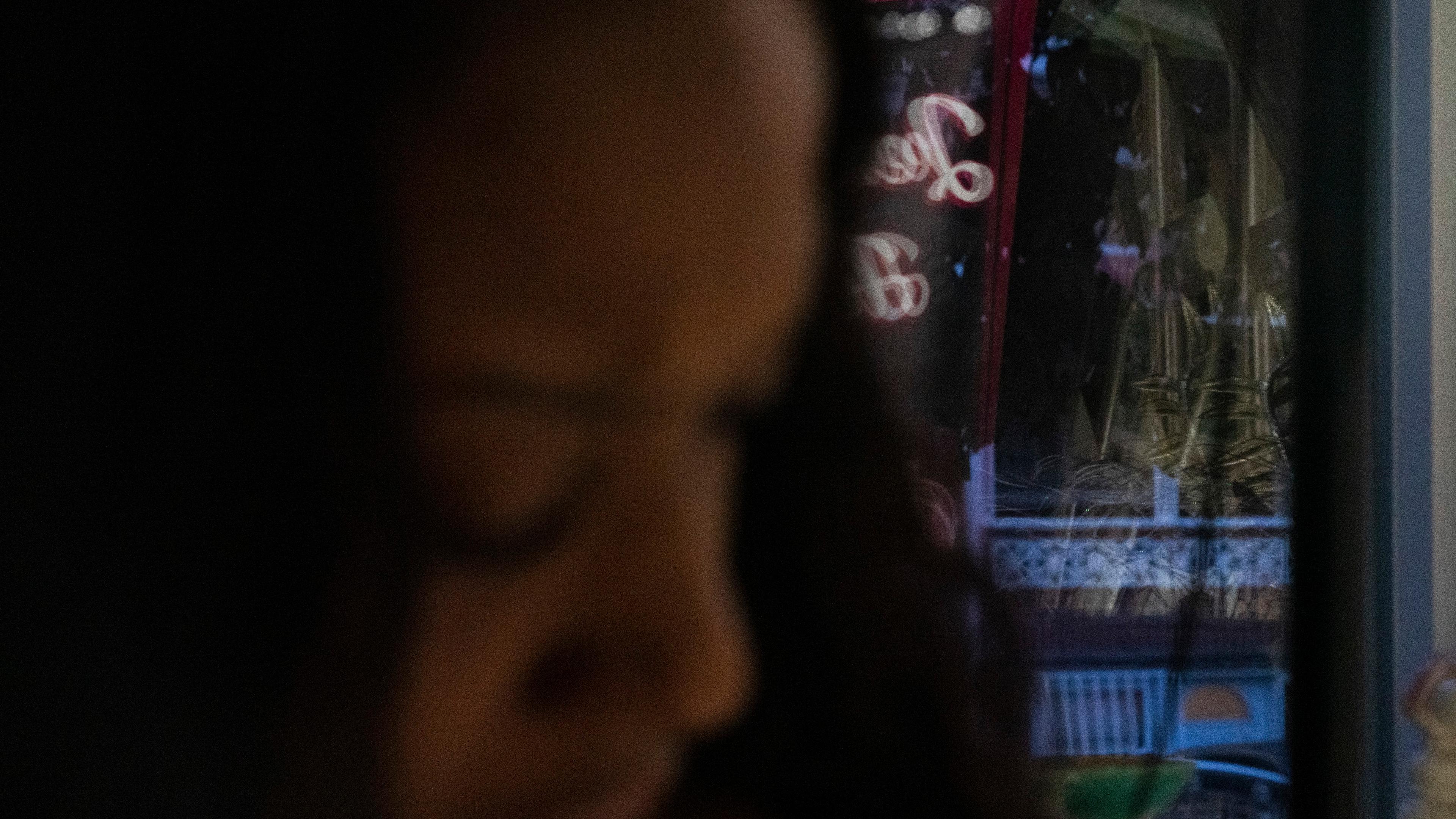 Blurry photo of a person with a colourful neon sign reflected in a window at night.