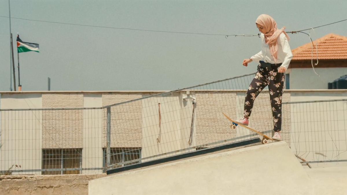 Photo of a young woman in a pink hijab skateboarding on a ramp, with a building and a flag visible in the background.