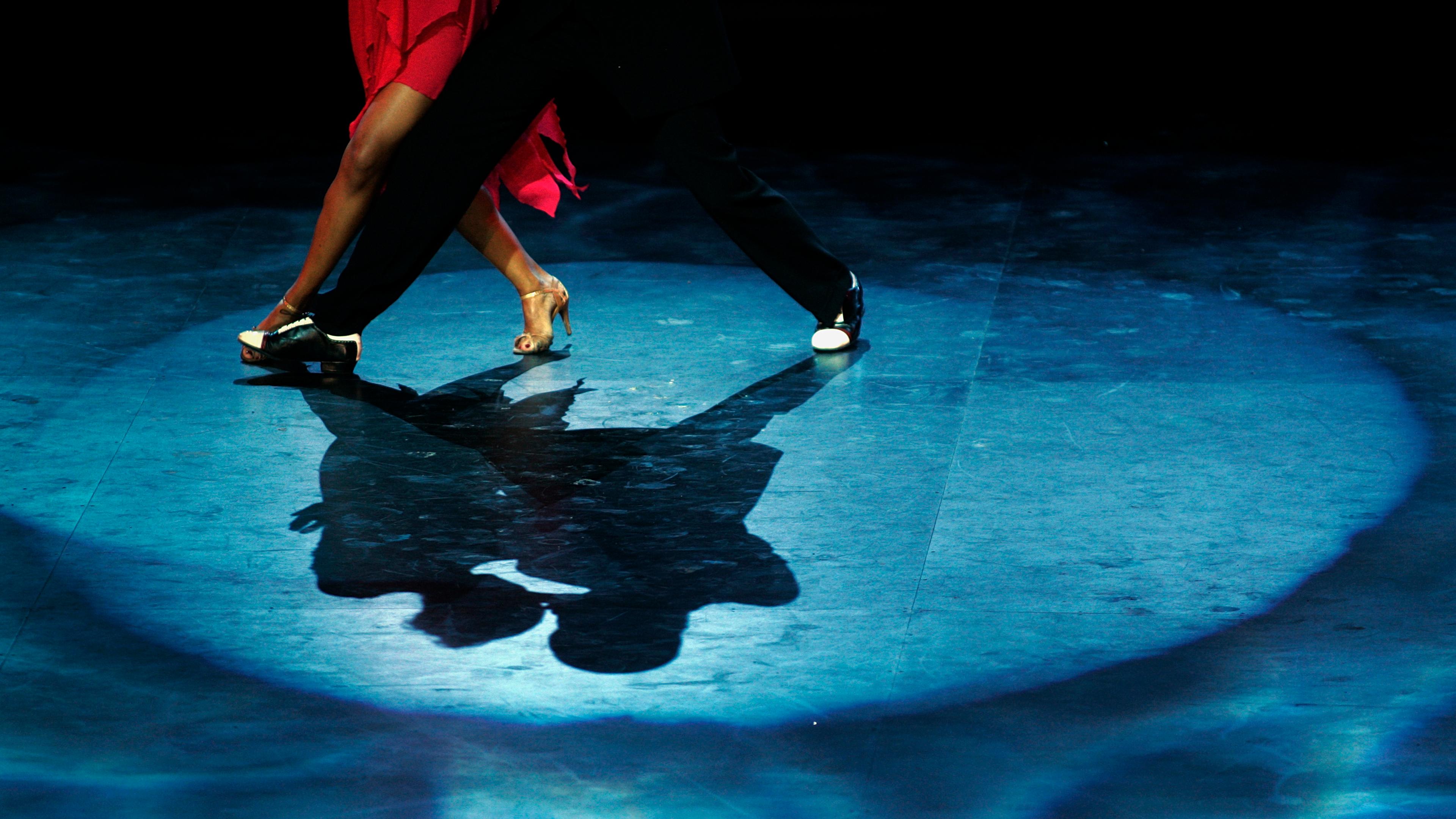 A couple dancing on stage, the photo focusing on their feet and shadows, with dramatic lighting and a blue spotlight.