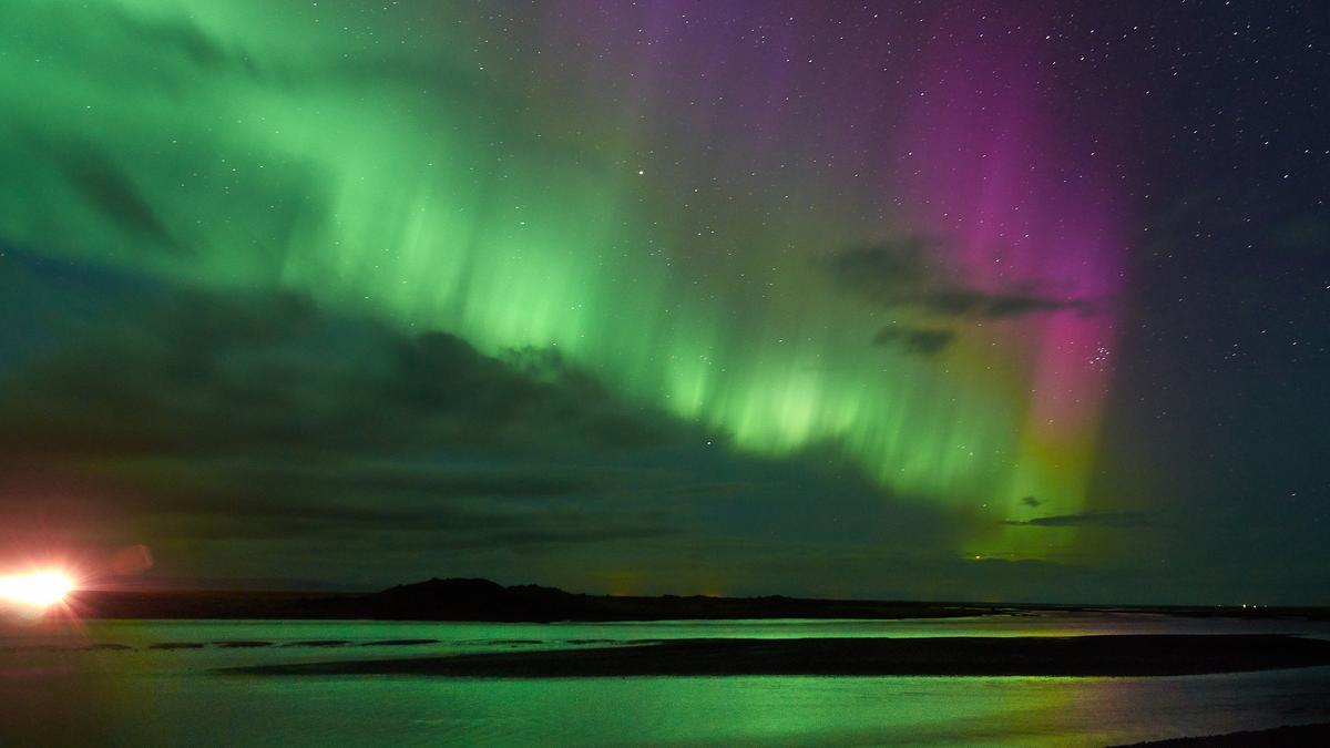 Photo of vibrant green and purple Northern Lights over a dark landscape with a bright glow on the horizon.
