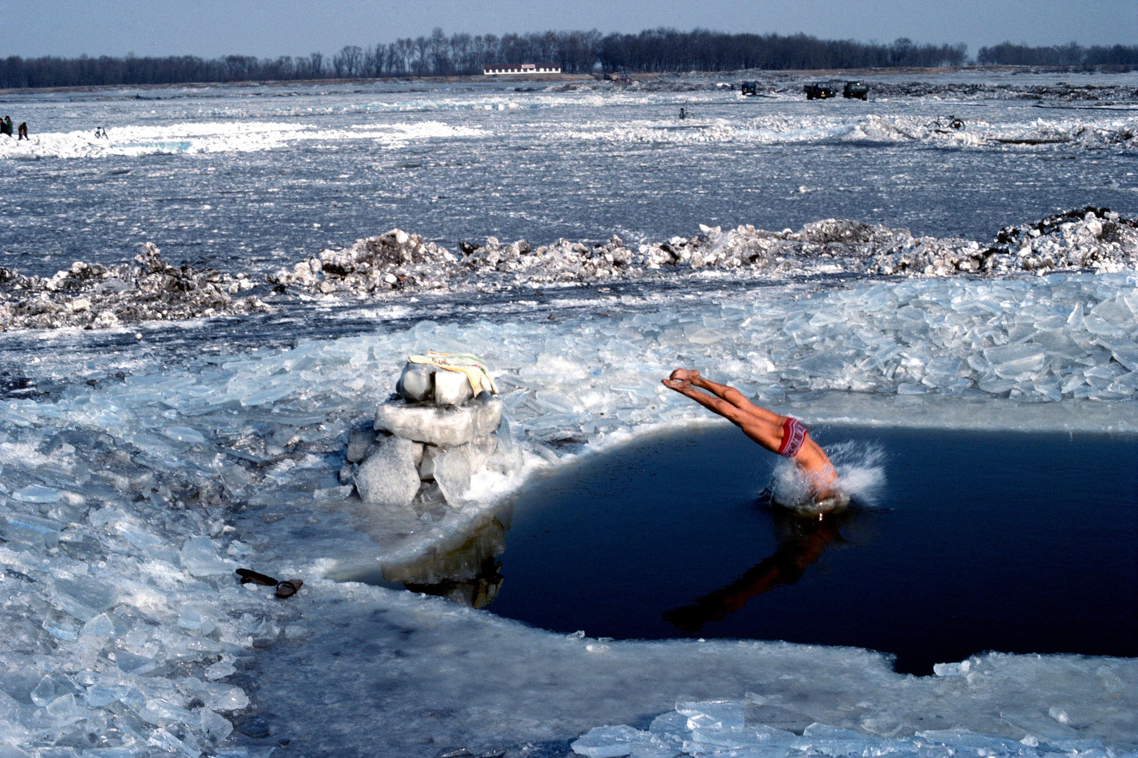 Photo of a person diving into an ice hole on a frozen river landscape with ice chunks and distant trees visible.