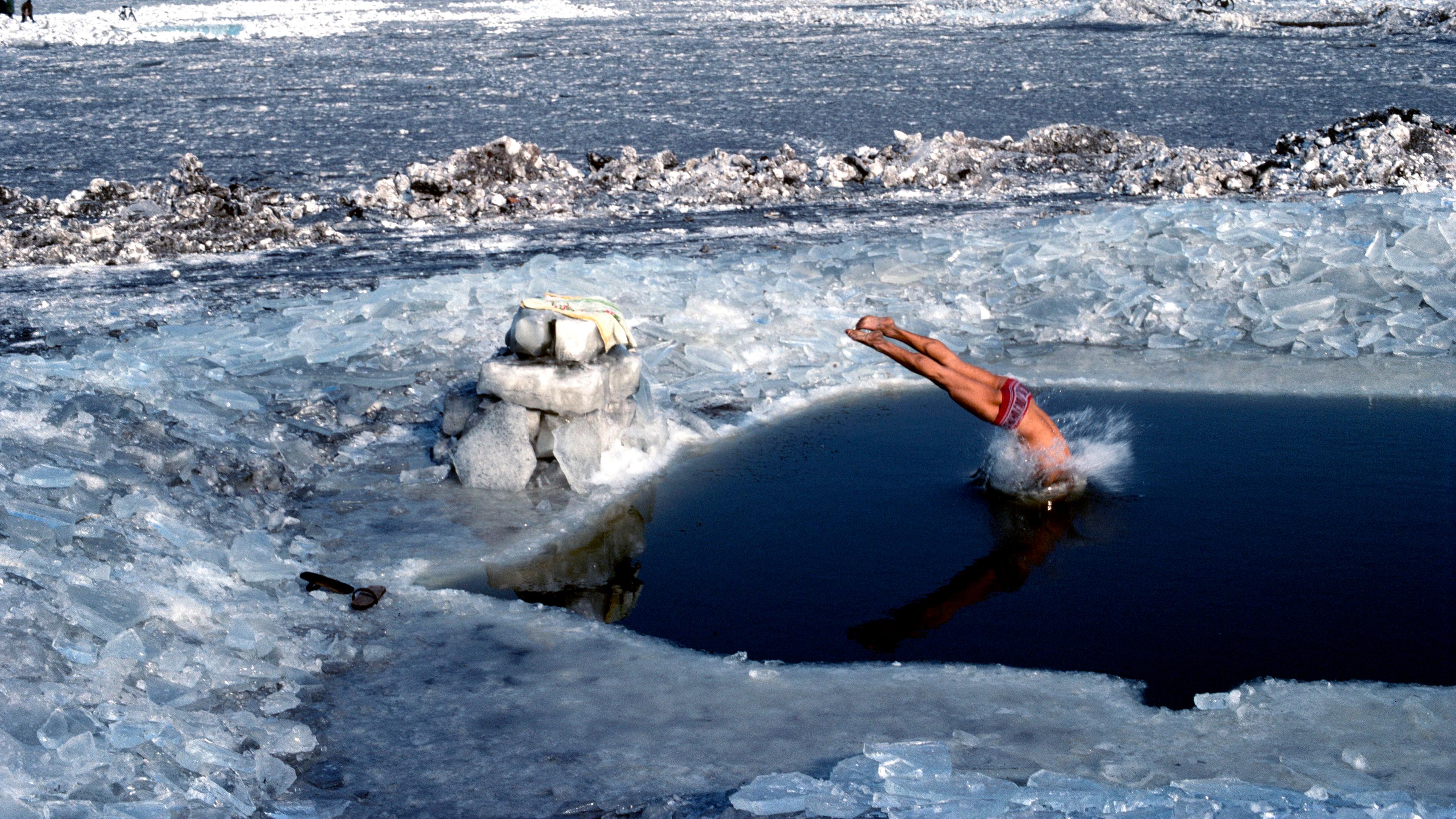 Photo of a person diving into an ice hole on a frozen river landscape with ice chunks and distant trees visible.