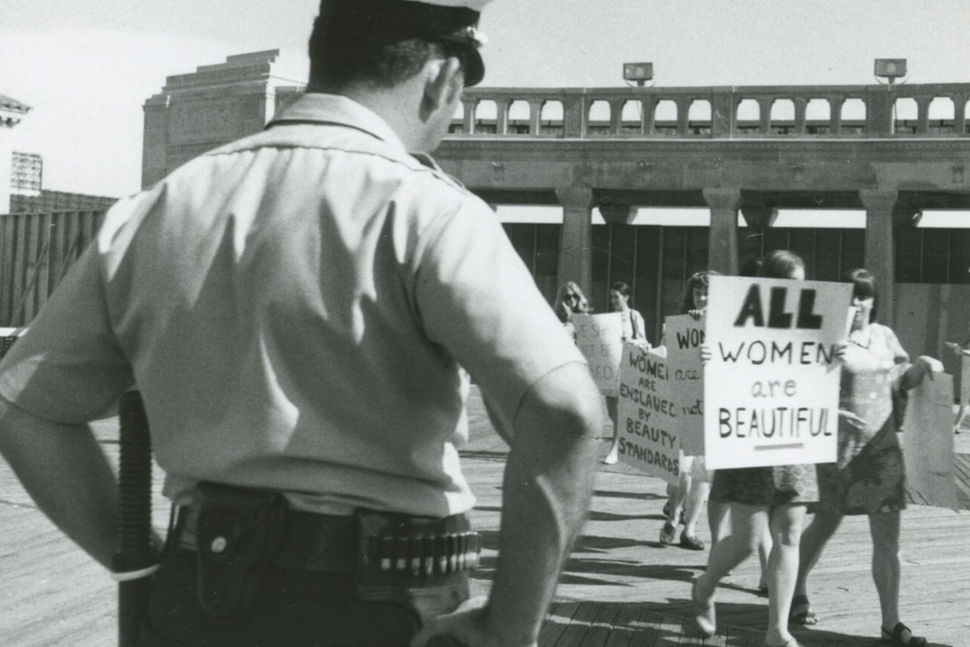 Black and white photo of a policeman observing a women’s protest with signs like “All Women Are Beautiful”