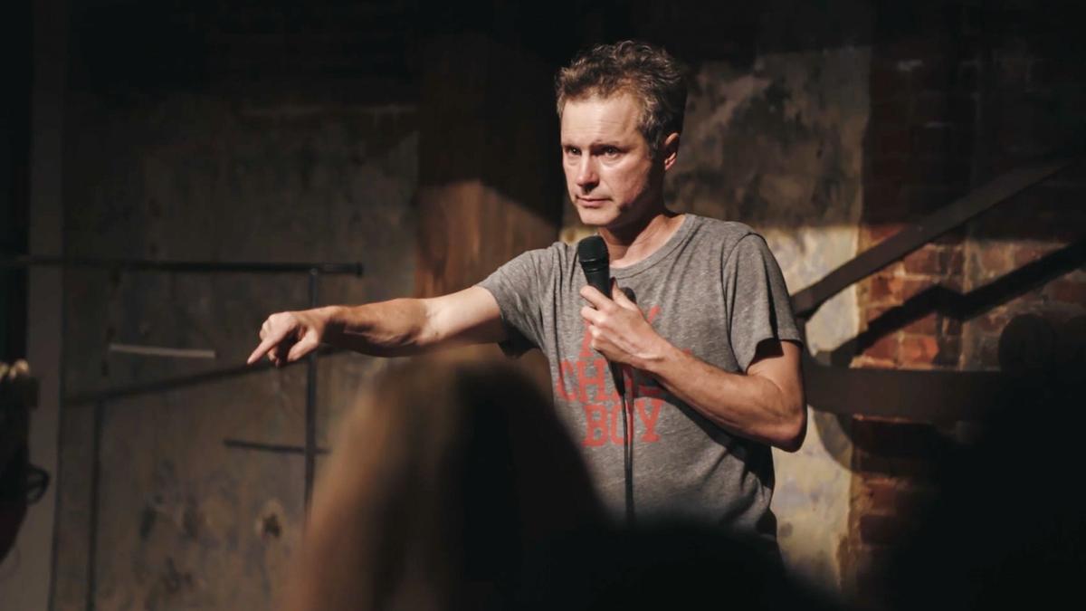 Photo of a man in a grey t-shirt holding a microphone pointing during a one-man show in a dimly lit venue.