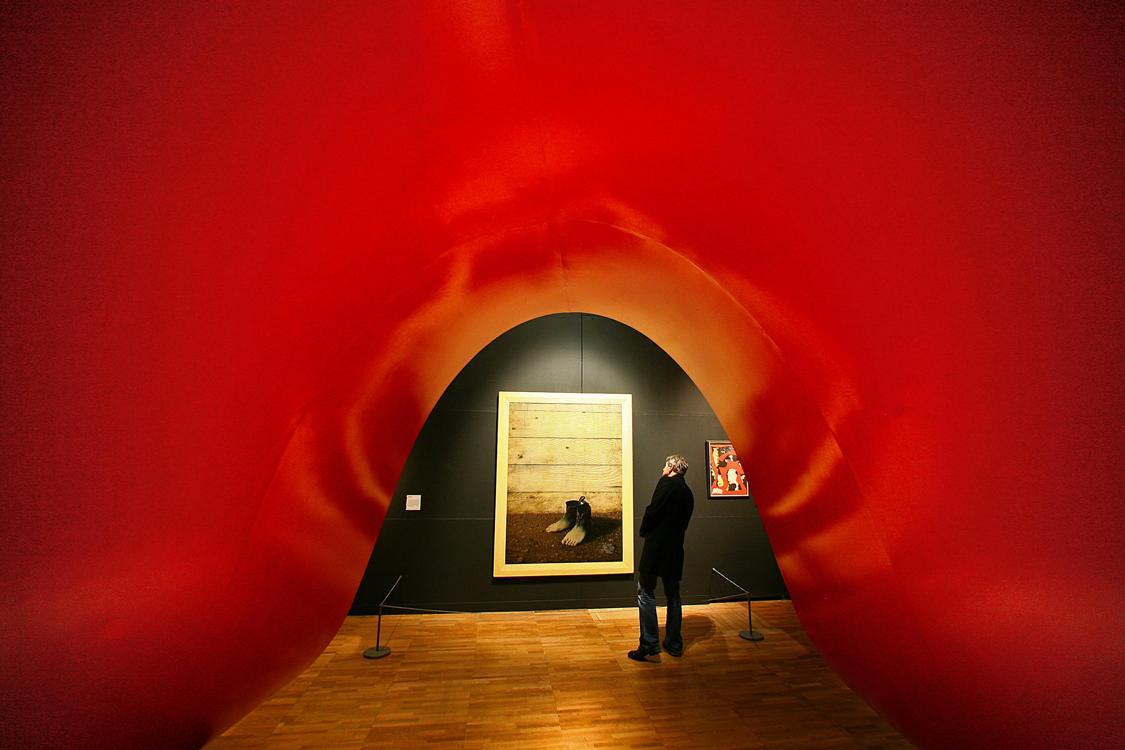 Photo of a person admiring art in a gallery framed by a large red sculpture, with a wooden floor and a dark wall background.