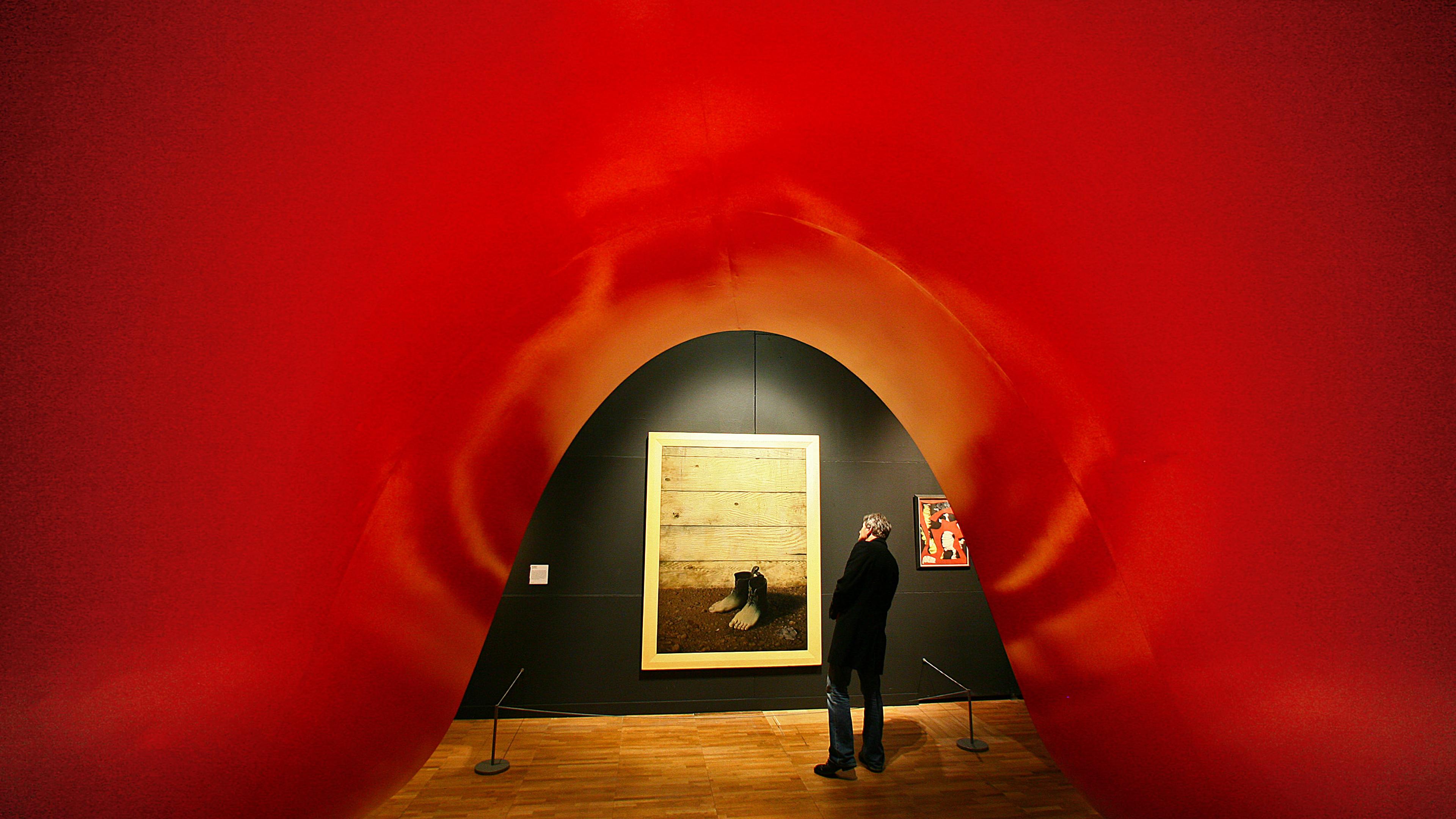 Photo of a person admiring art in a gallery framed by a large red sculpture, with a wooden floor and a dark wall background.