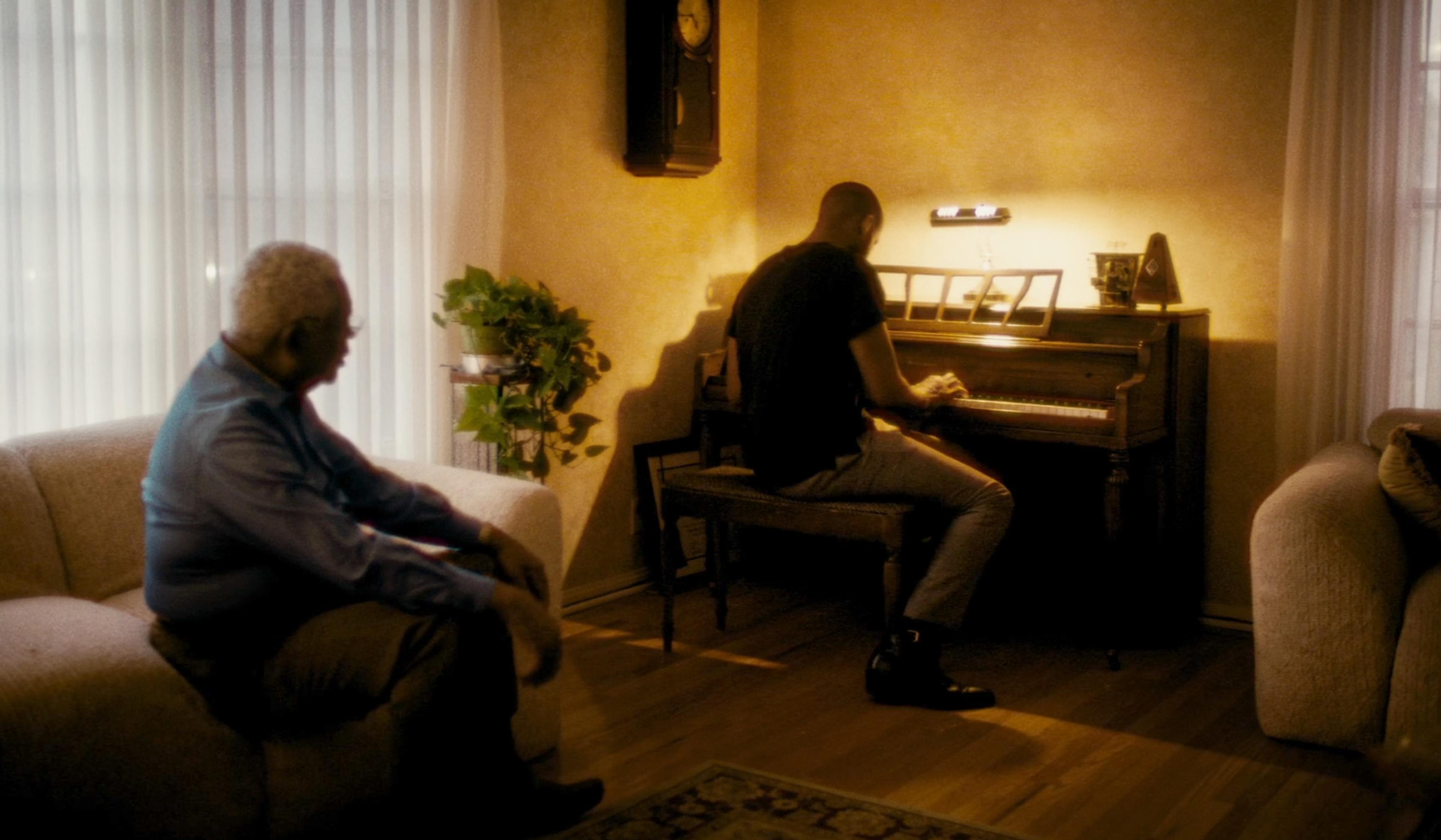 A man plays piano in a living room while another man watches from a sofa, illuminated by warm light.