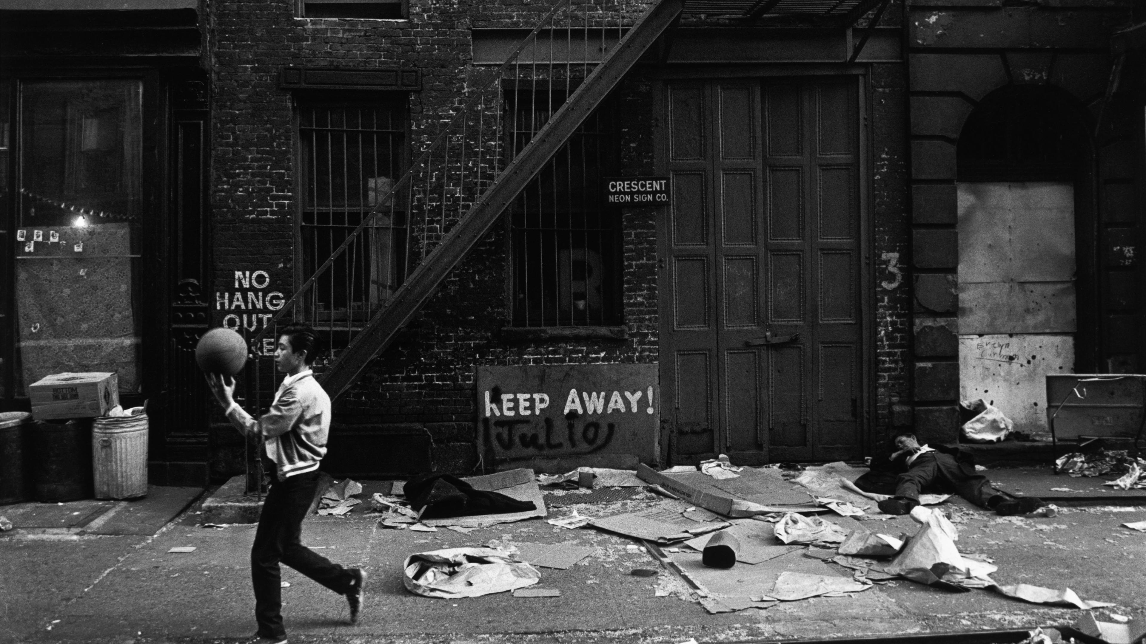 Black and white photo of a man with a ball on a littered street, graffiti and a fire escape visible on a brick building.