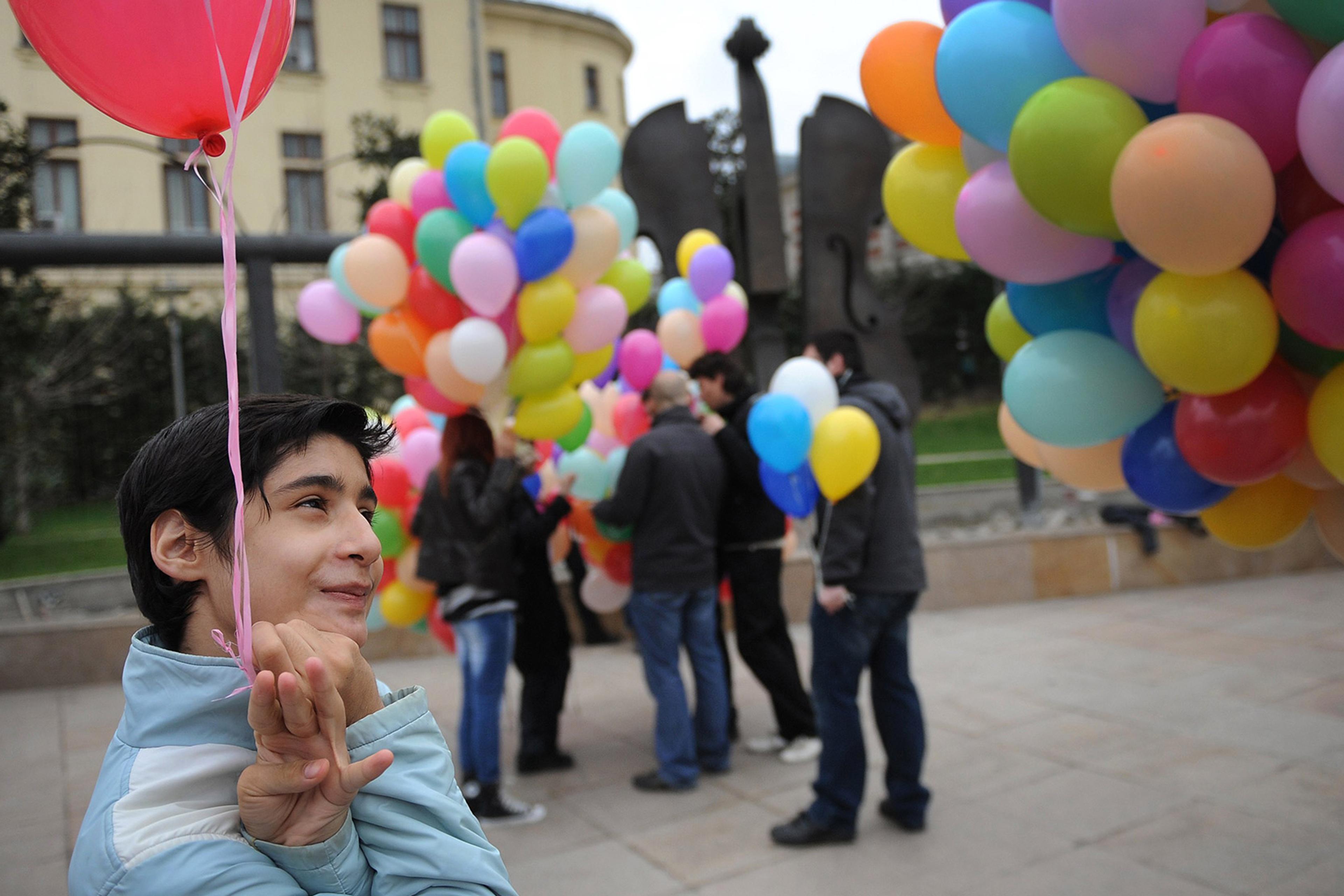 A smiling neurodiverse child holds a red balloon. In the background people hold balloons of various colours