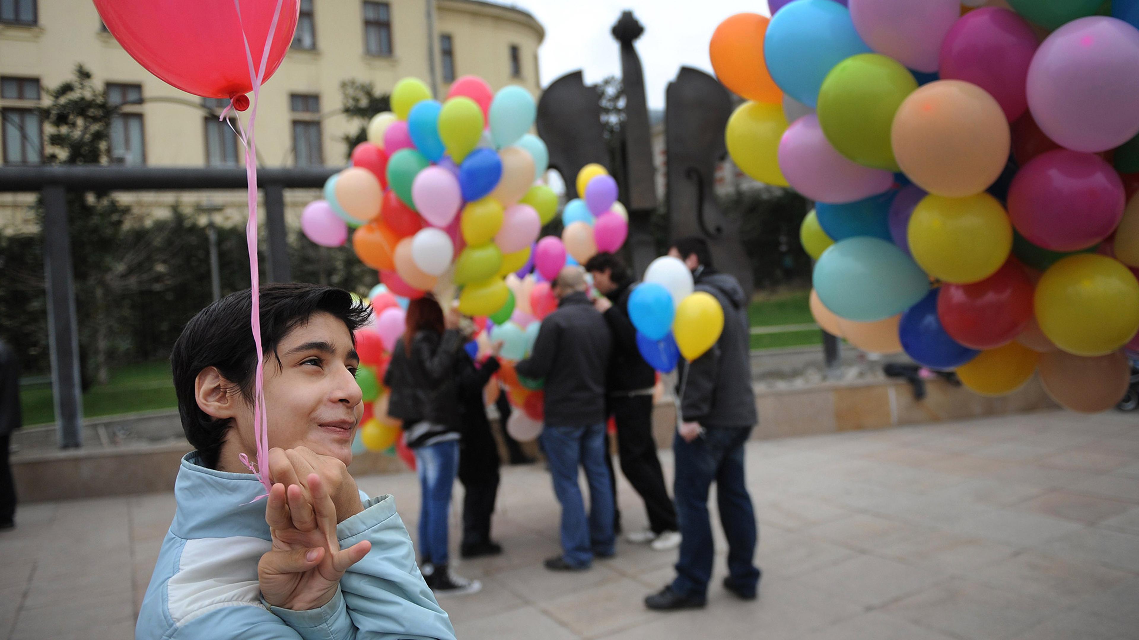 A smiling neurodiverse child holds a red balloon. In the background people hold balloons of various colours