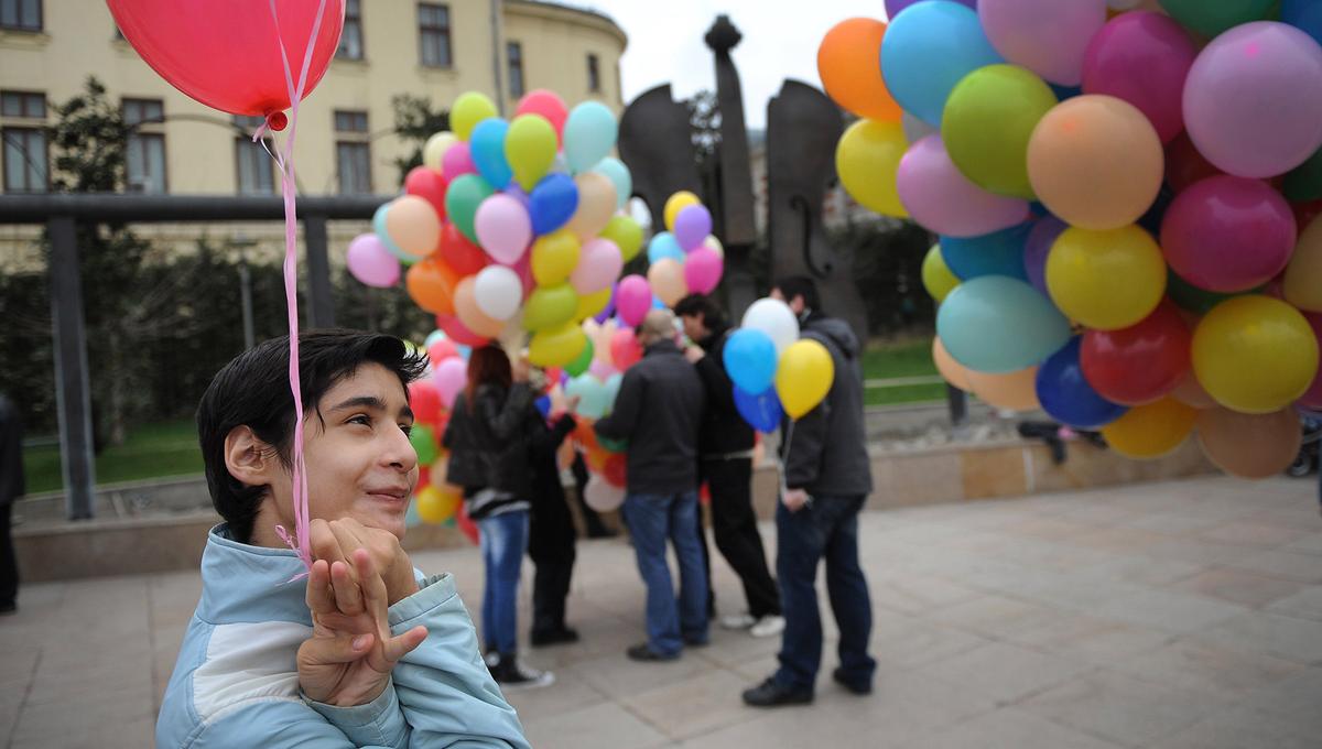 A smiling neurodiverse child holds a red balloon. In the background people hold balloons of various colours