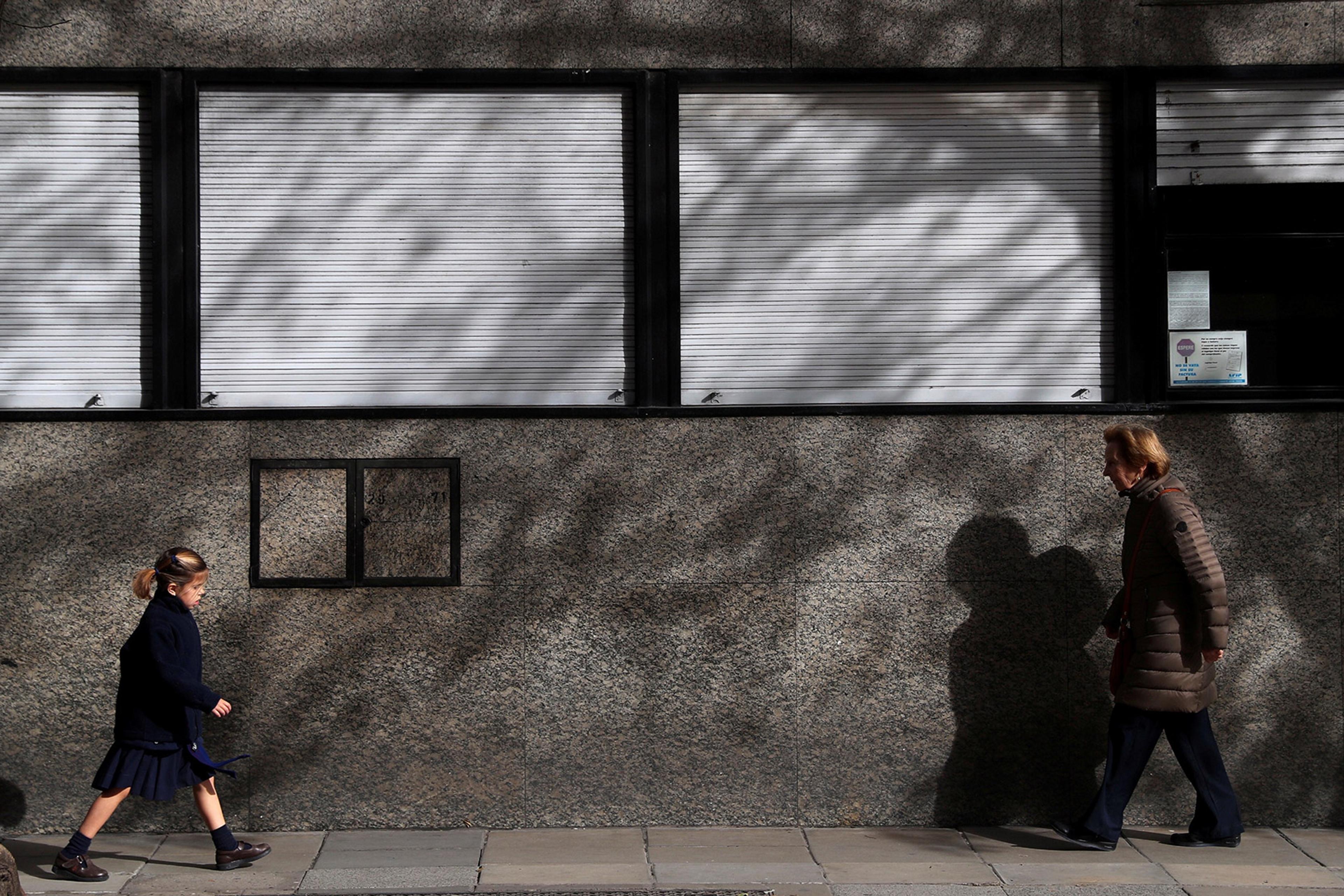 A girl walks towards a woman on a pavement with a grey building in the background, casting shadows from a tree.