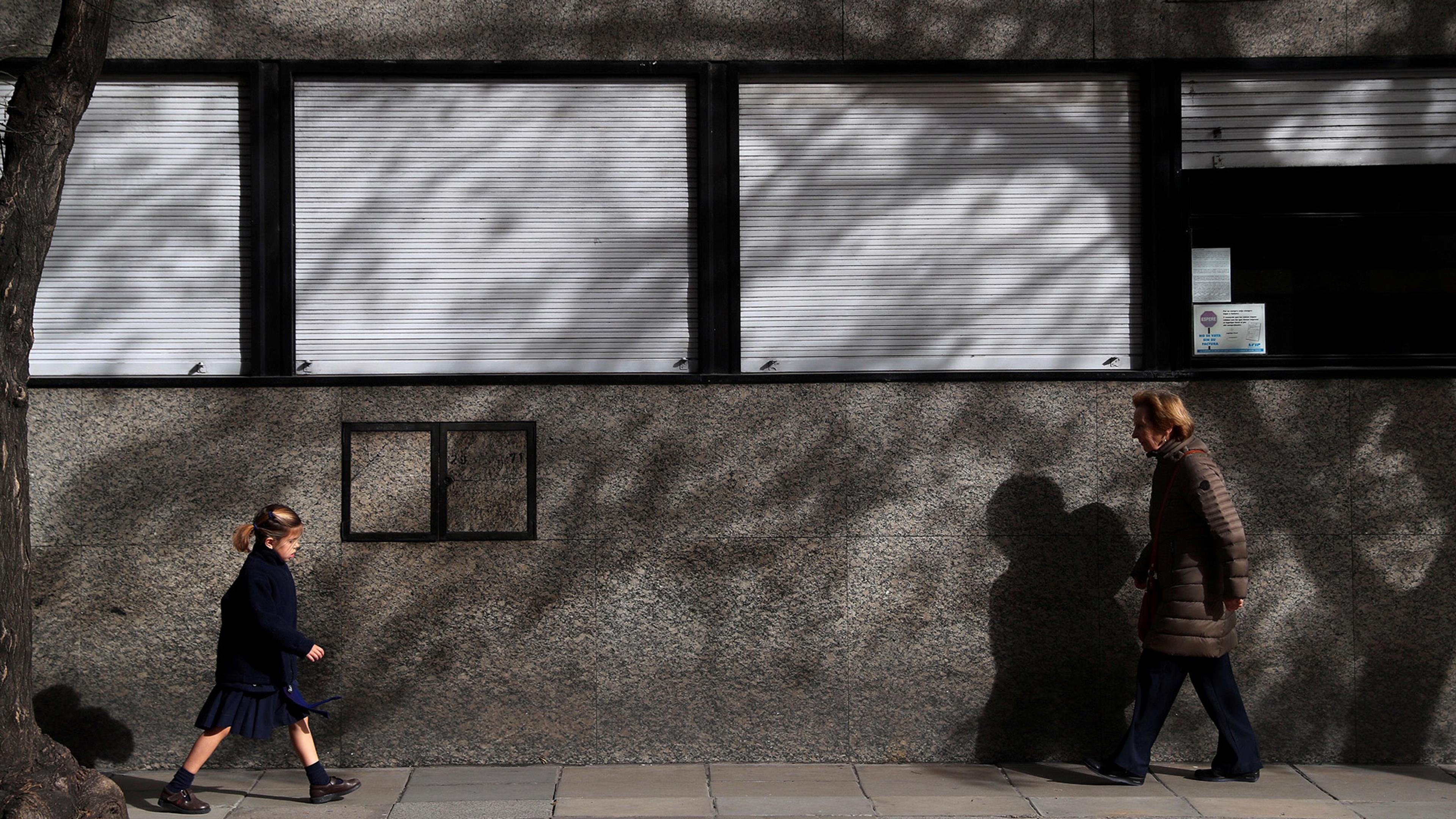 A girl walks towards a woman on a pavement with a grey building in the background, casting shadows from a tree.