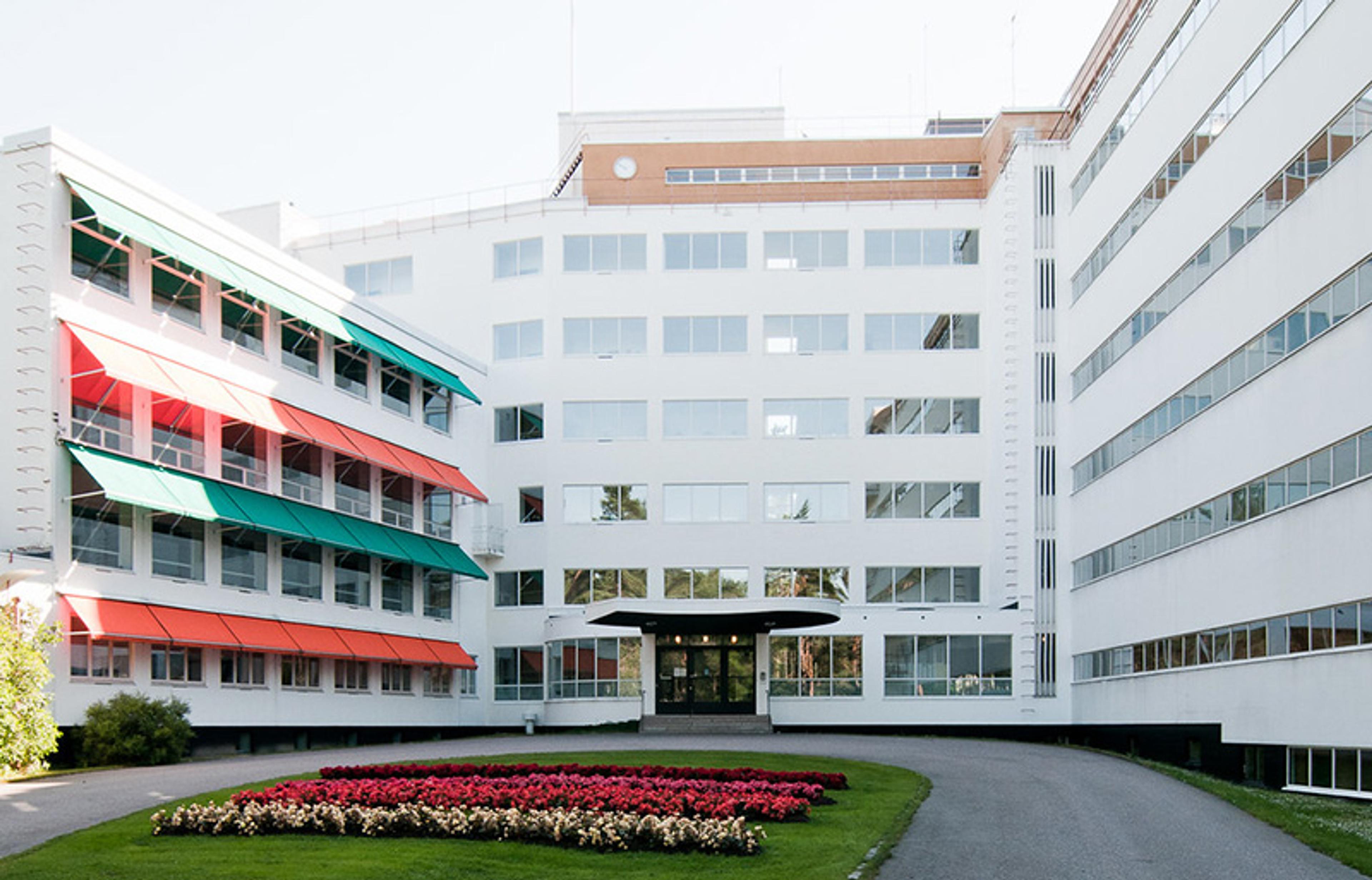 A modern white building with multiple windows and colourful awnings, a pathway and flowerbed leading to the entrance.