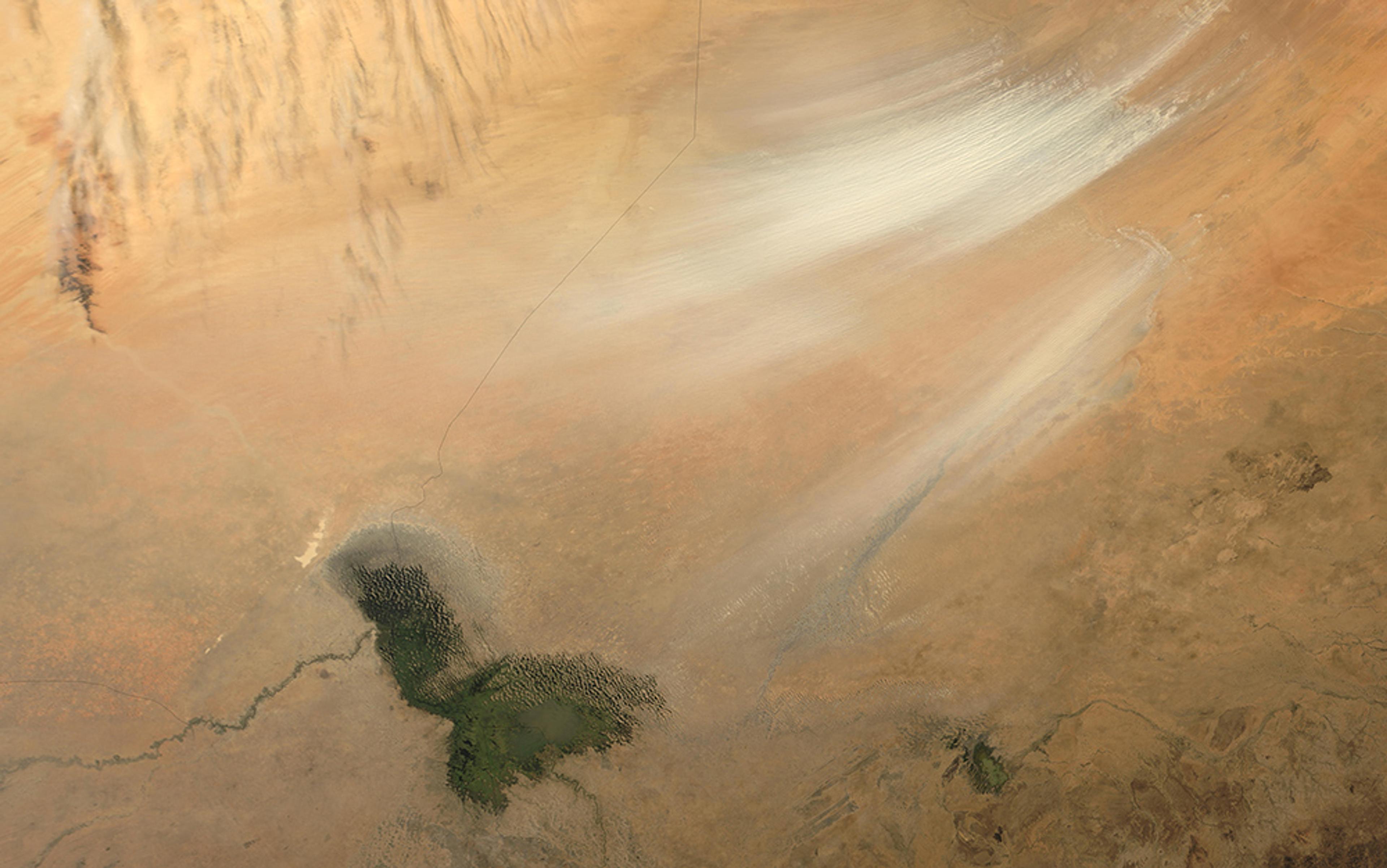 Satellite image of a desert region with sandy terrain, a patch of vegetation, and wispy clouds above.
