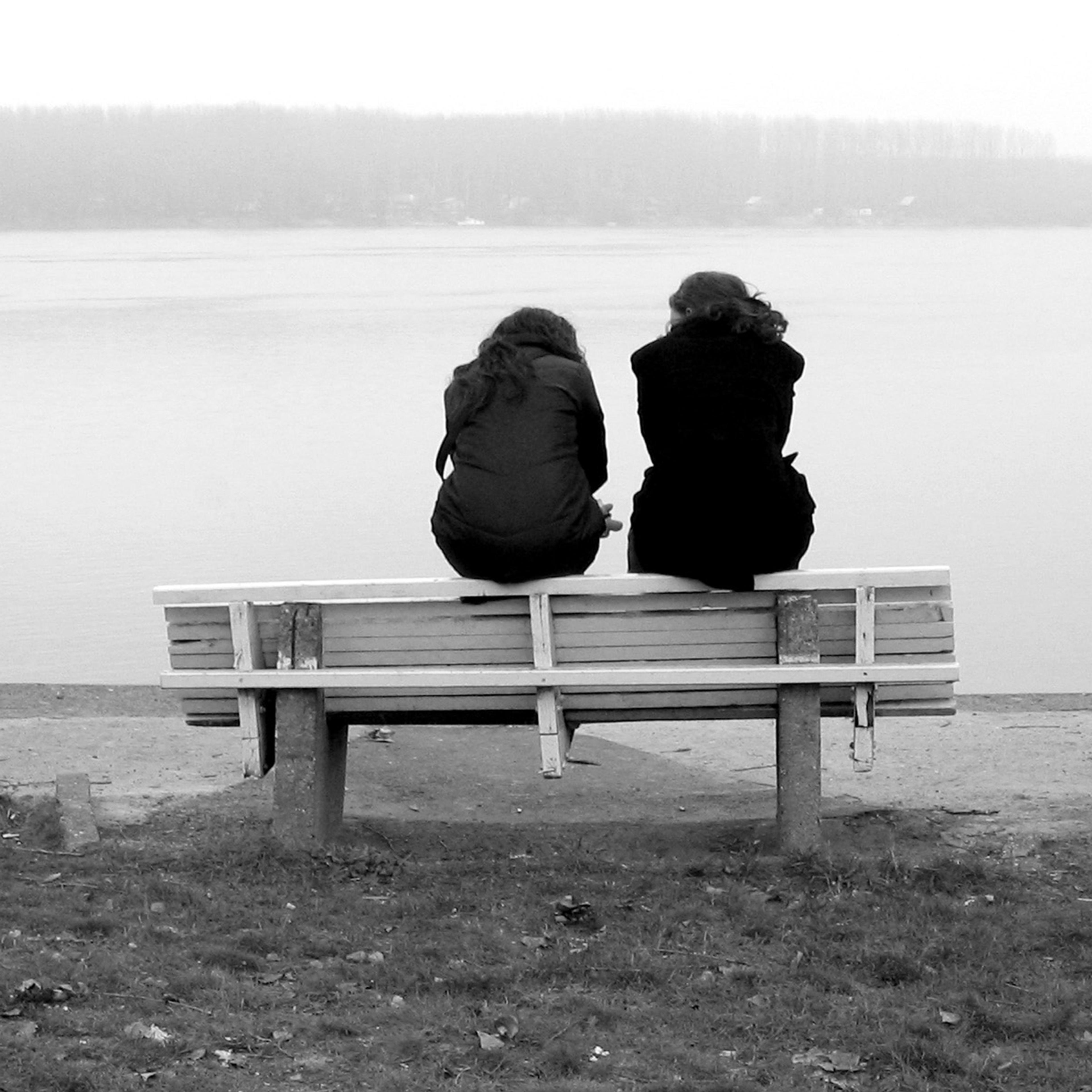 Black and white photo of two people sitting on a bench by the water, viewed from behind, with trees visible on a distant shoreline.