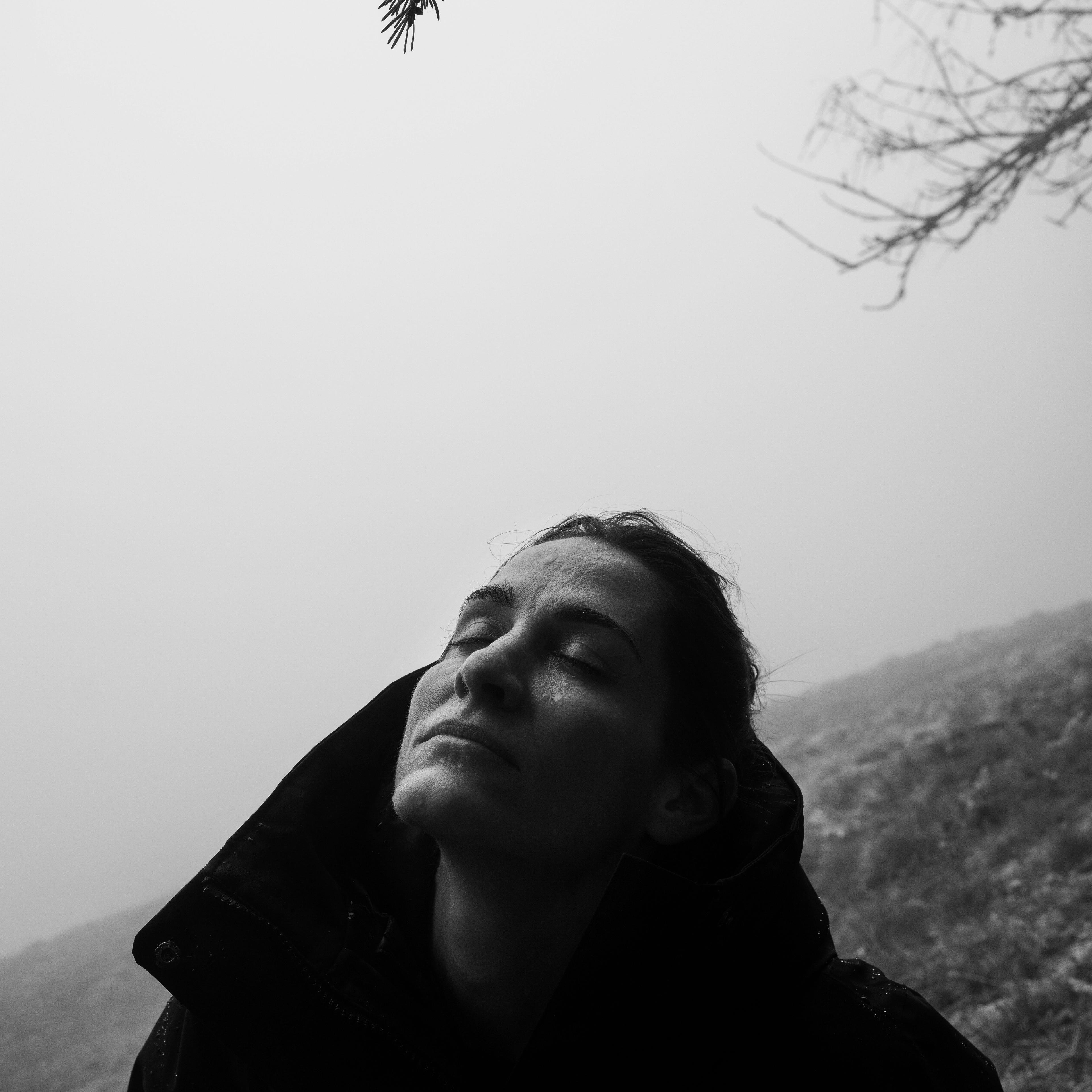 Black and white photo of a person wearing a jacket, eyes closed, with a foggy forest background and tree branches above.