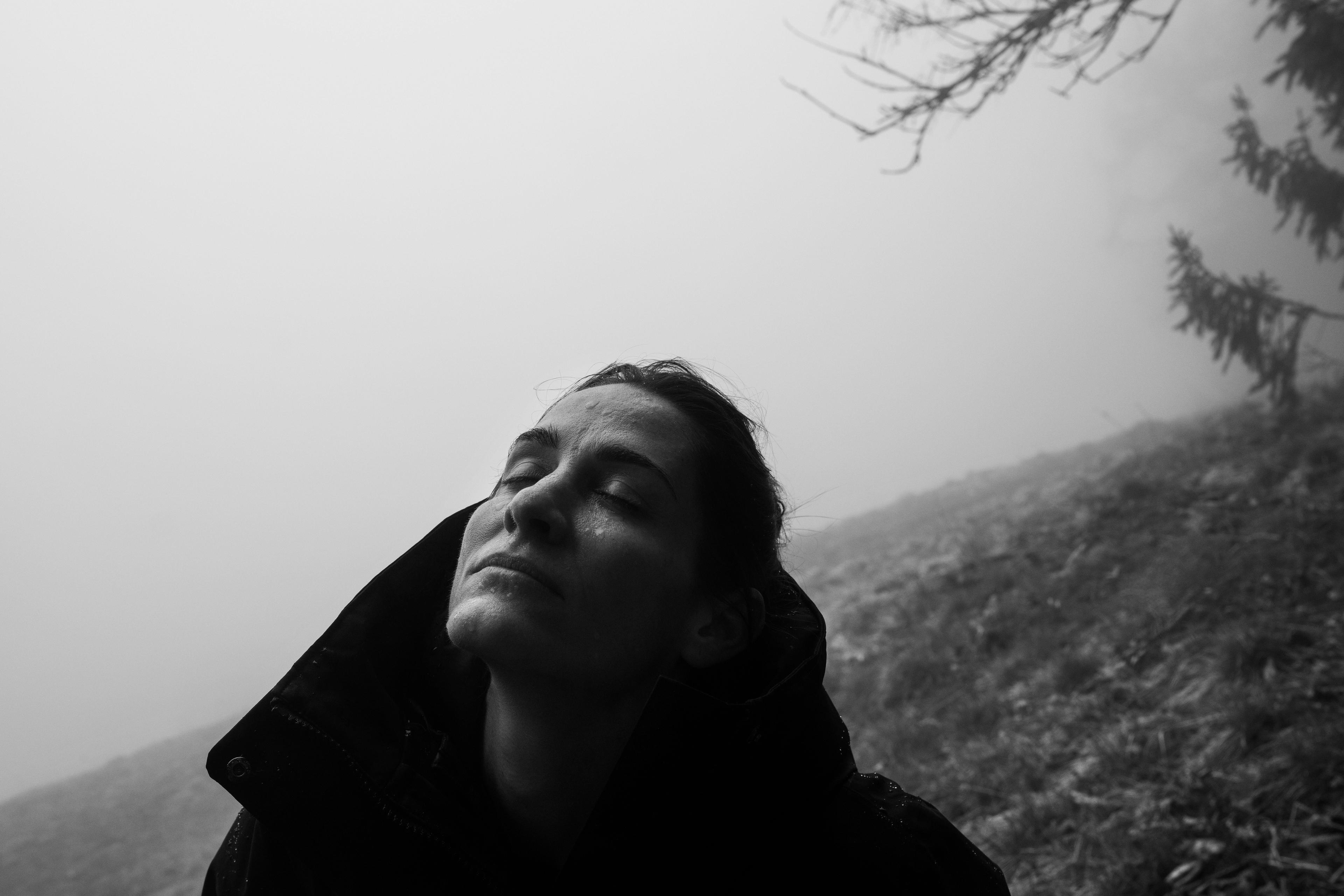 Black and white photo of a person wearing a jacket, eyes closed, with a foggy forest background and tree branches above.