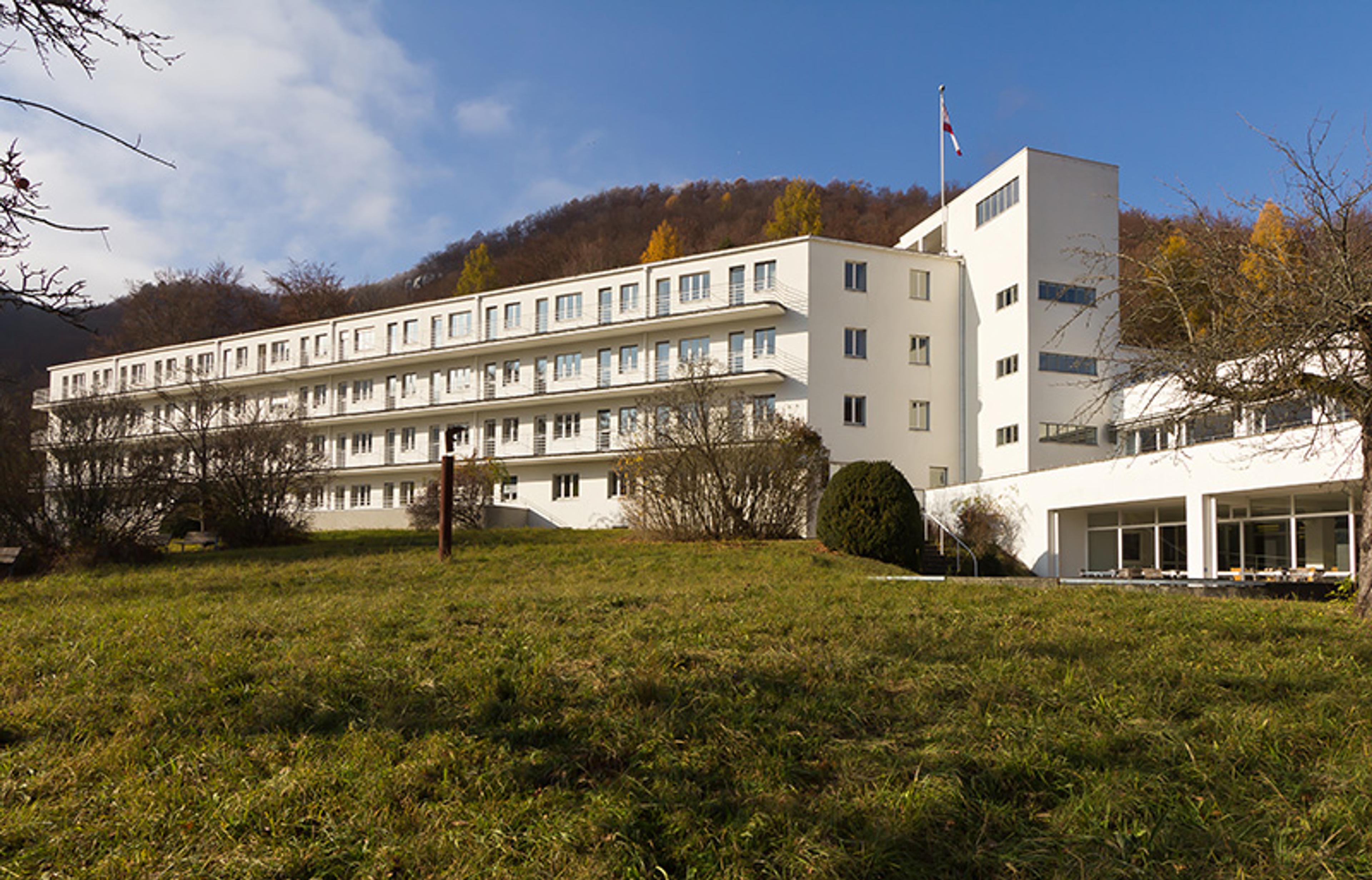 A large white multi-story building on a grassy hill with trees in the background under a partly cloudy sky.