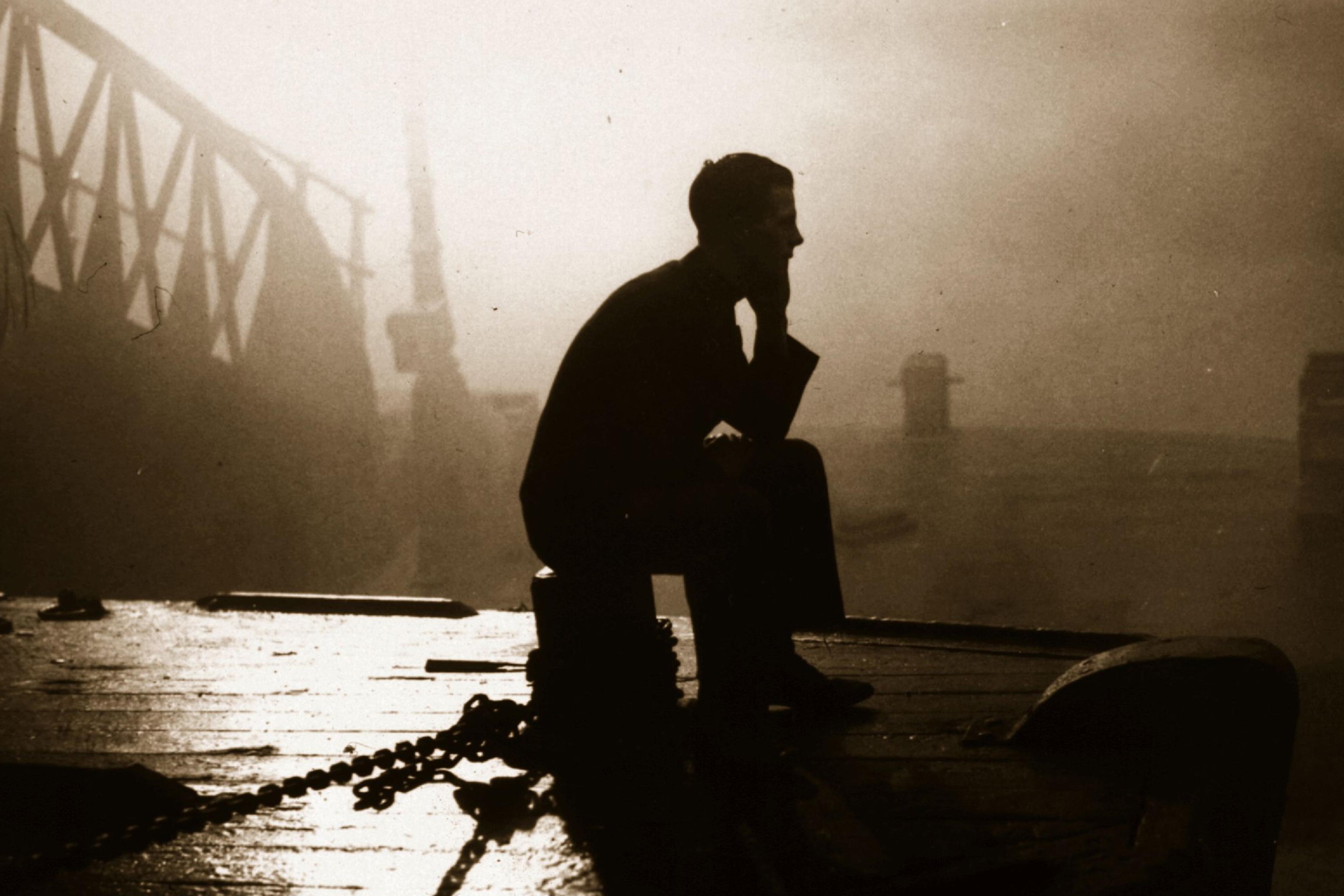 Silhouette of a person sitting on a docked boat at sunset with a bridge and mist in the background, photograph.