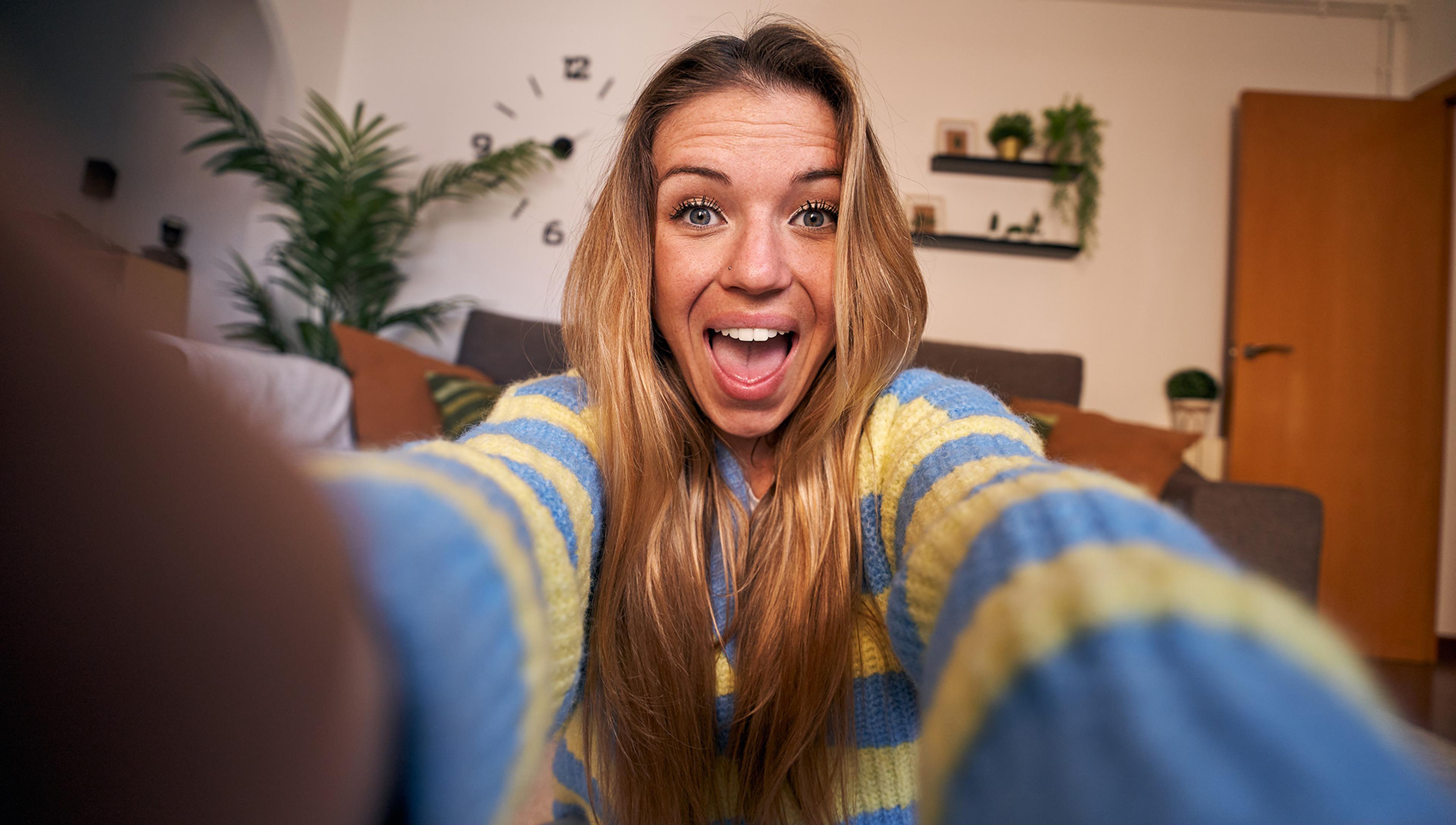 A woman with long blonde hair and a striped blue and yellow jumper takes a selfie, smiling broadly. Behind her, a living room is visible with a large potted plant, a grey couch, and wall shelves with small plants and decor items. A clock is fixed on the wall in the background.