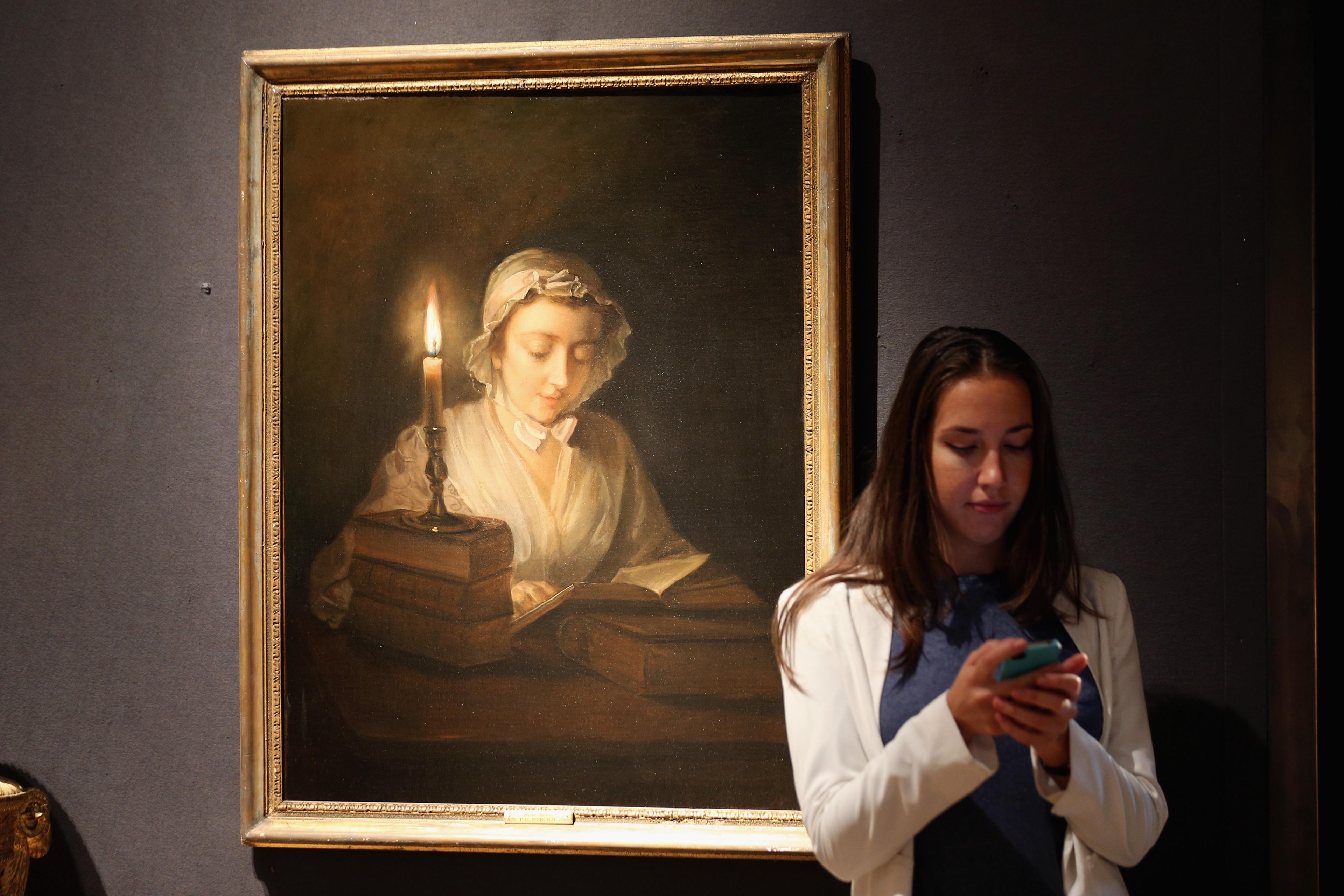 A woman using a phone stands beside a painting of a girl reading by candlelight.