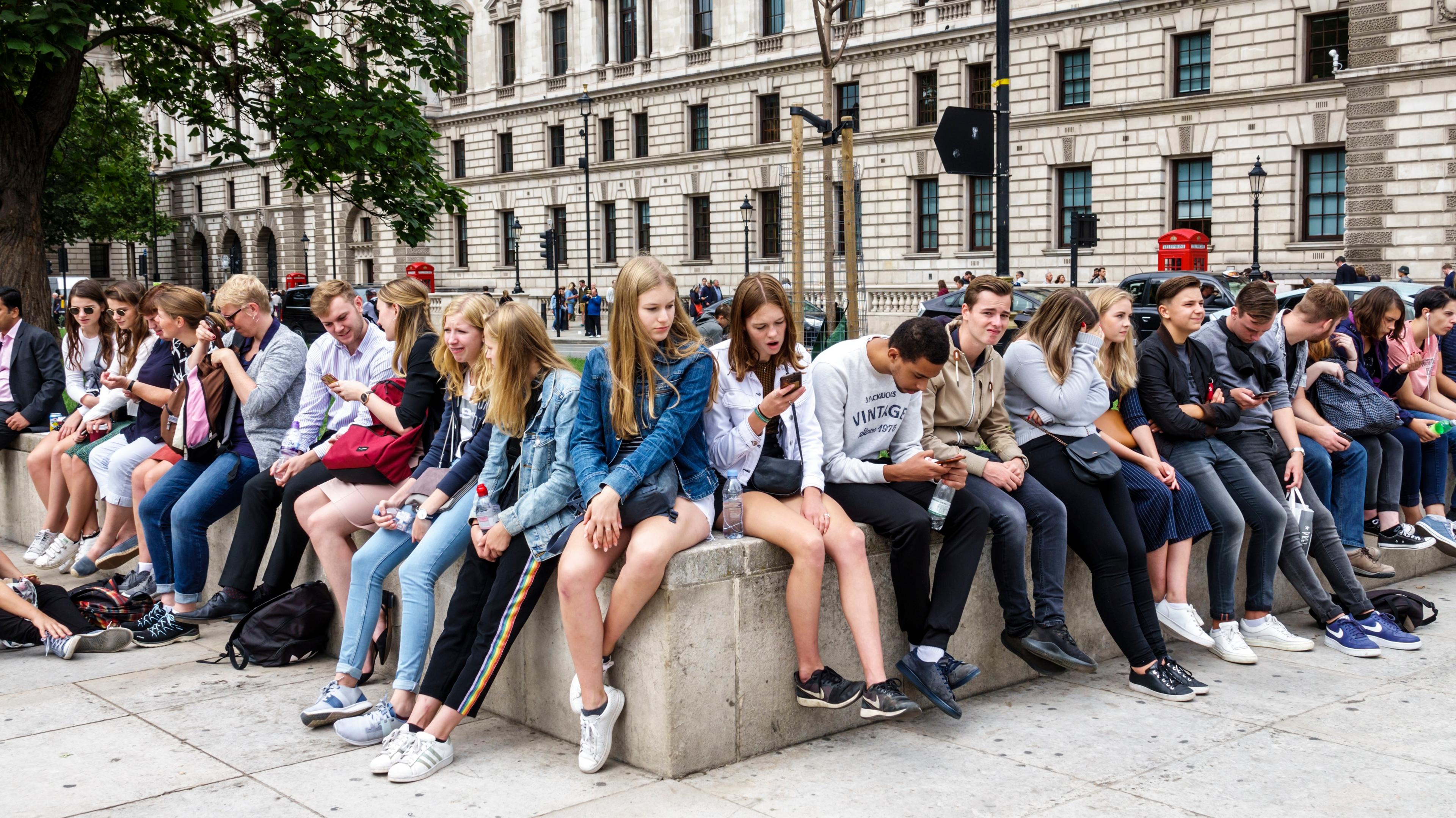 Photo of young people sitting on a stone ledge outside a historic building, some using phones, with trees and red phone boxes.