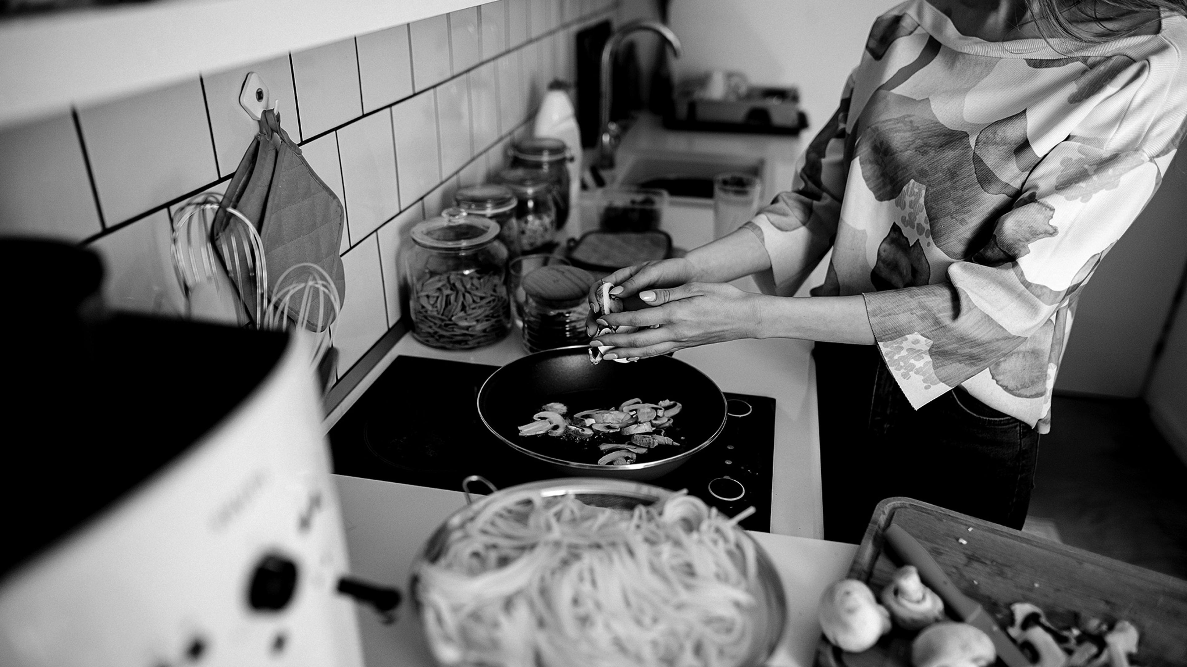 Black and white photo of a woman cooking in a kitchen with pasta and mushrooms on the counter.