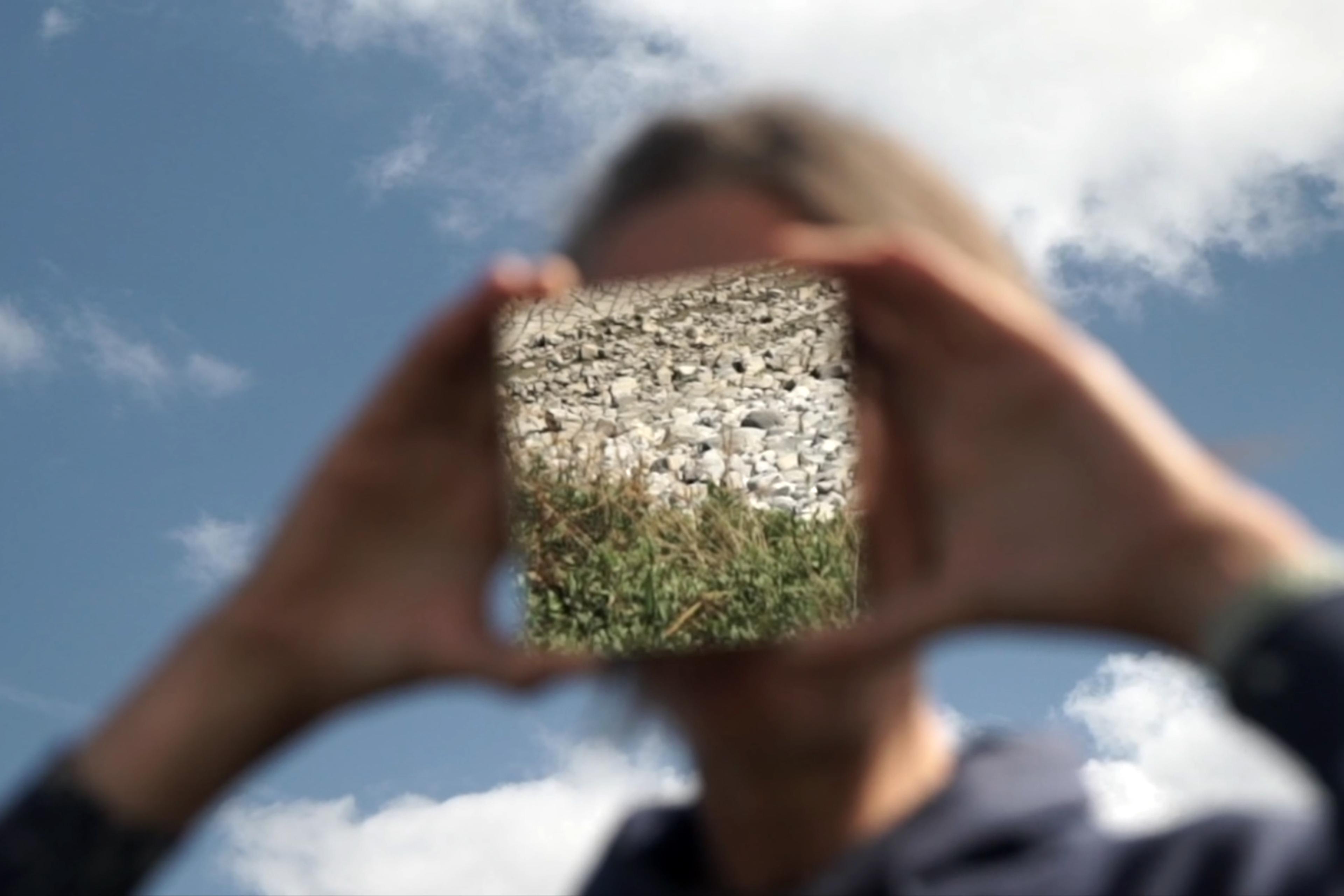 A woman holds a square mirror that blocks her face; it reflects rocks and grass. There is blue sky and clouds in the background. The only element in focus is the reflection on the mirror.