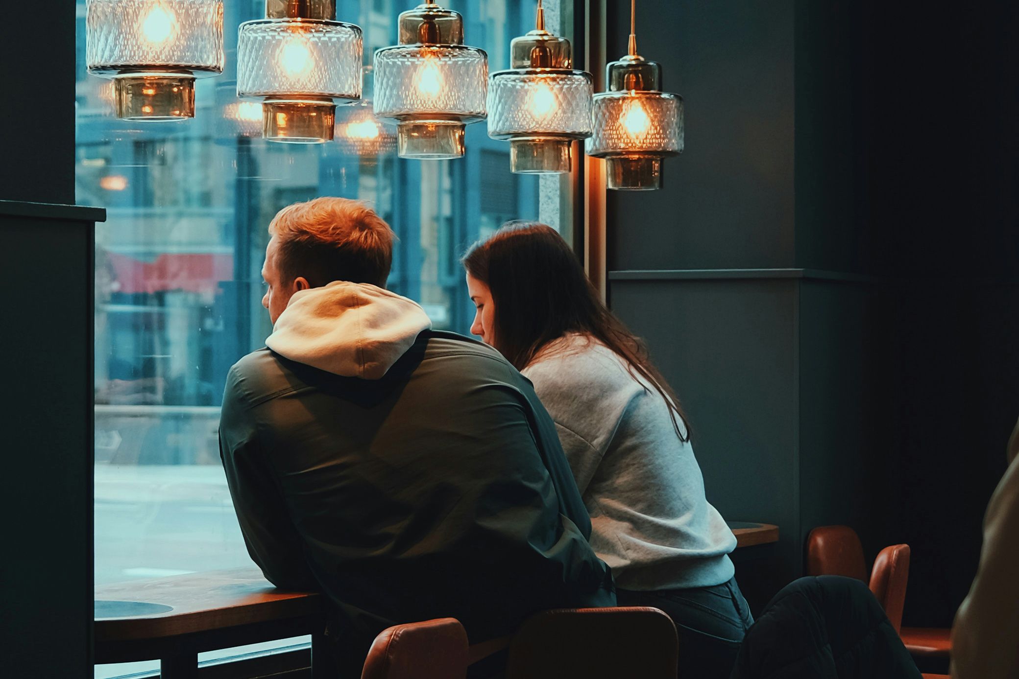 Two people sitting at a café table by a window under hanging lights, looking outside at a cityscape.